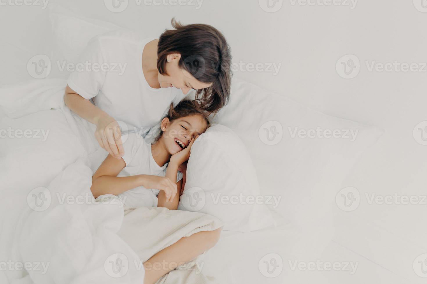 Overjoyed mum and daughter have fun together in bedroom, pose under soft blanket. Happy small girl spends leisure time with mother, has toothy smile, expresses happiness. Children and parents photo