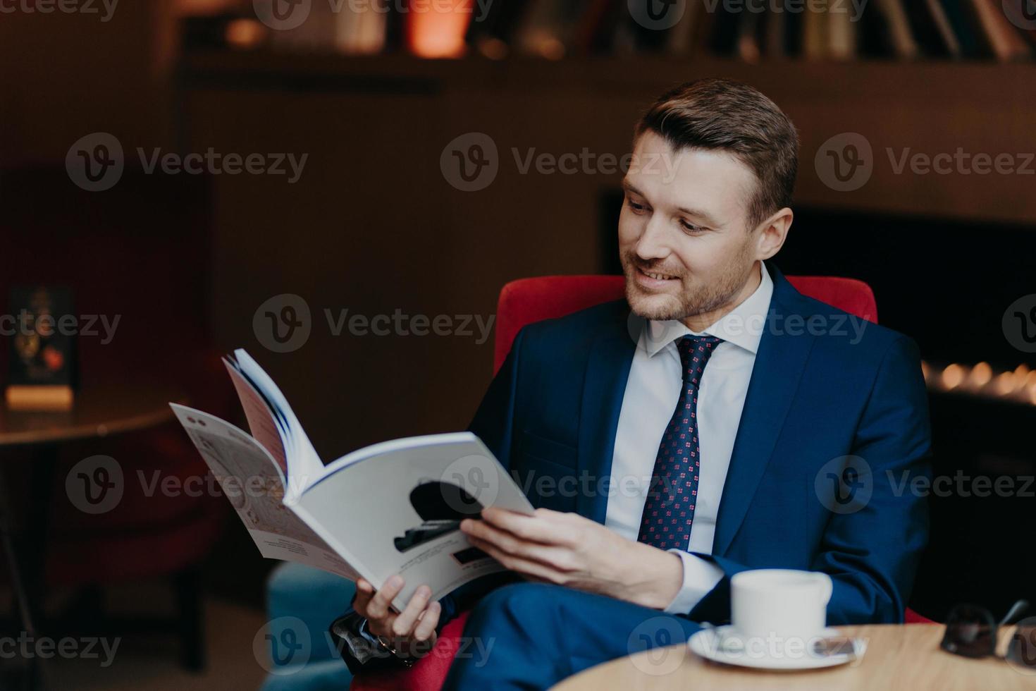 Portrait of happy male comapny owner reads magazine, rests in cozy coffee shop, has happy expression, has break after business meeting, dressed in formal clothes. People, rest and leisure concept photo