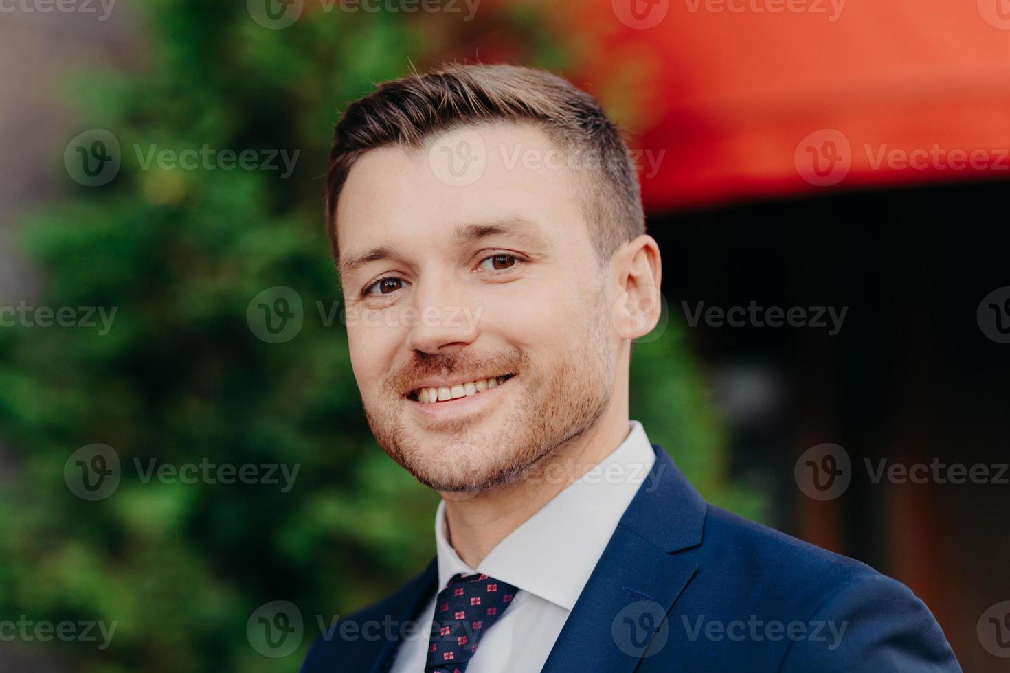 Close up portrait of handsome attractive male with gentle smile, stubble, wears elegant clothing, looks directly into camera, poses outdoor, satisfied with getting success. Male entrepreneur photo
