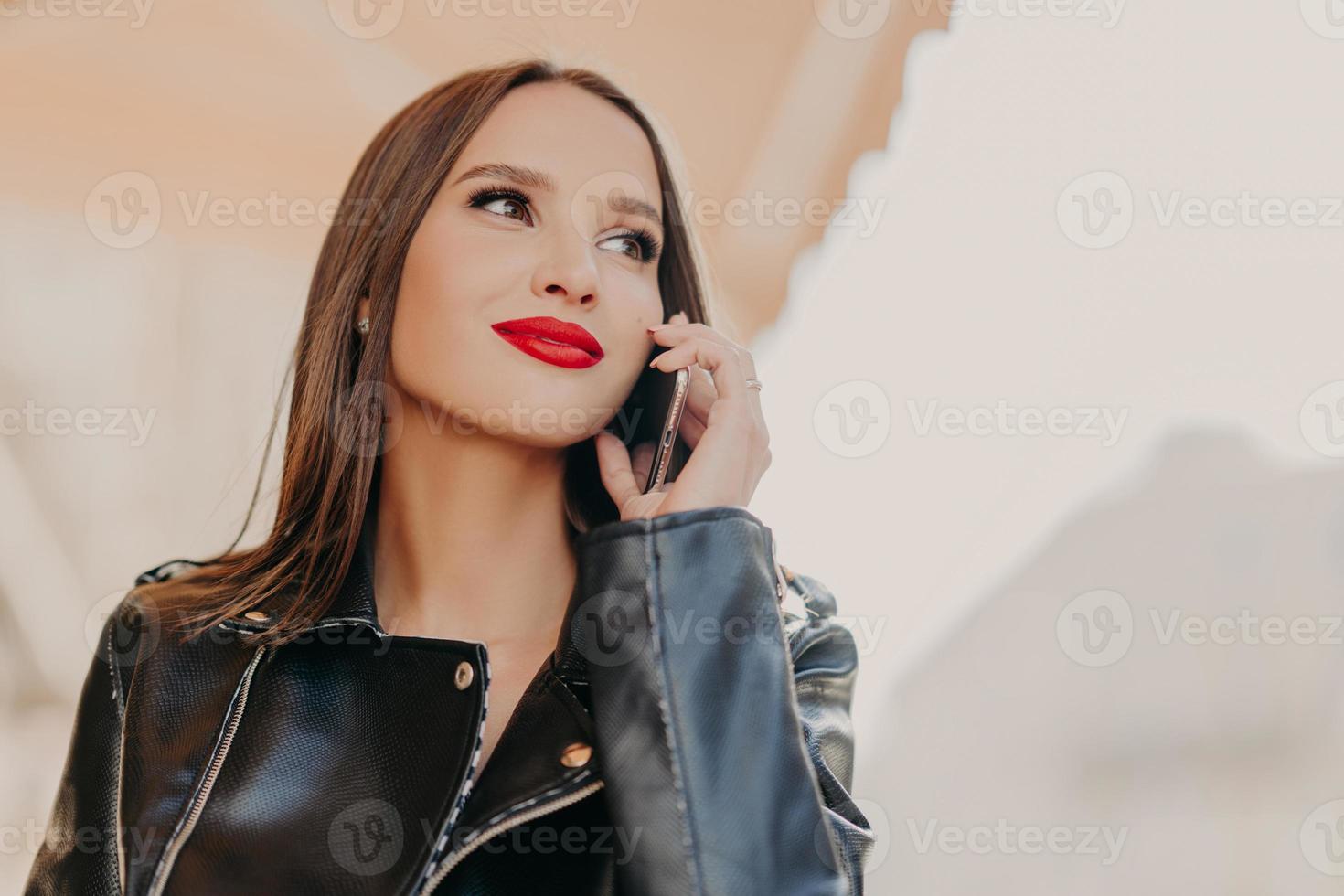Headshot of pleasant looking dreamy woman focused aside, has telephone conversation, wears leather coat, notices someone into distance, poses over blurred background. Good roaming connection photo