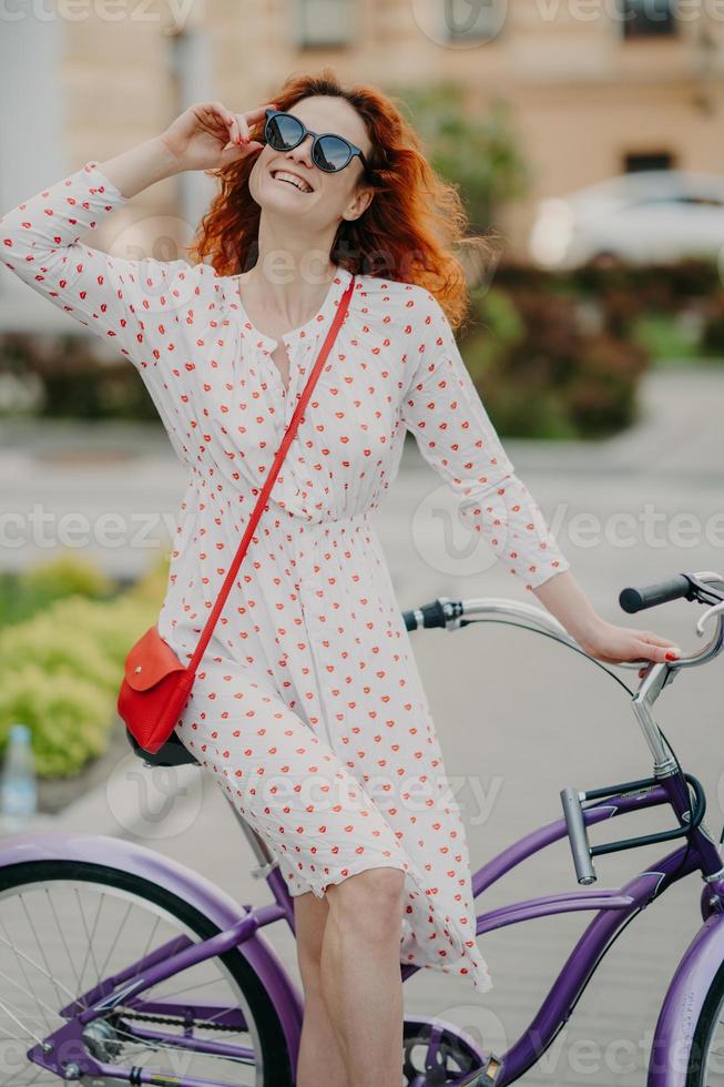 Active cheerful woman rides bicycle in city, wears stylish sunglasses, has toothy charming smile, dressed in summer dress, enjoys sunny day, spends free time actively. People, transport and lifestyle photo