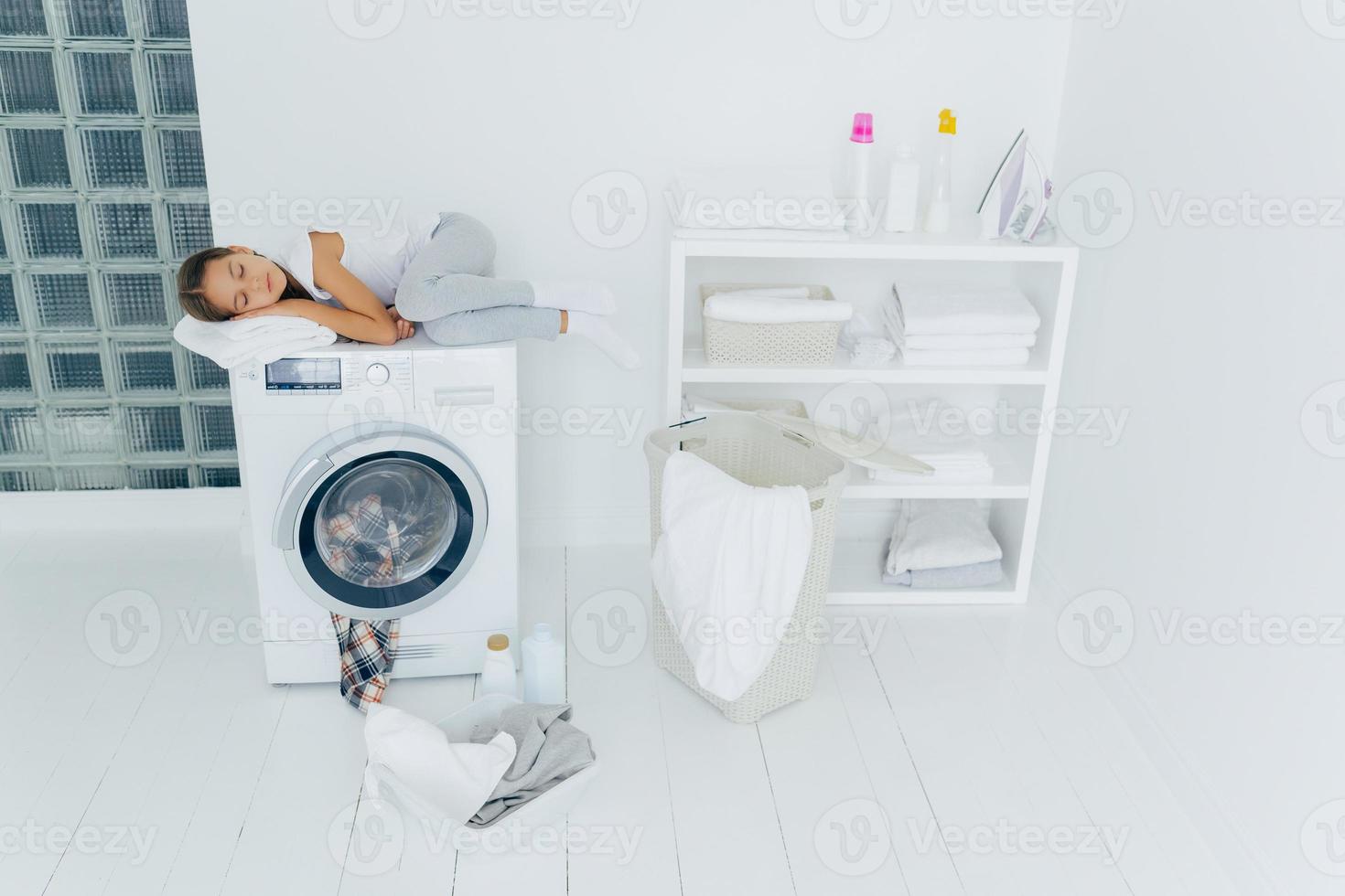 Shot of tired small kid sleeps on washing machine, uses white soft towel as pillow, has pleasant dreams, basket and basin on white floor, feels fatigue after loading clothes in washer, being at home photo