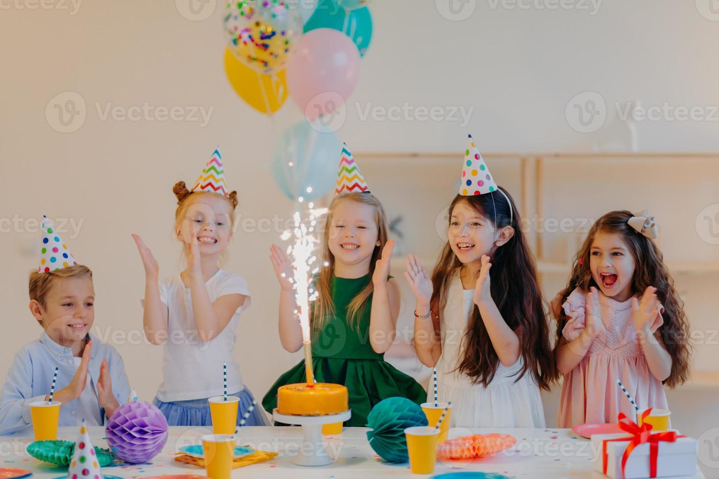Overjoyed children friends clap hands and looks at big sparkle on cake, stand near festive table with present, cups and holiday attributes, have fun together, celebrate birthday photo