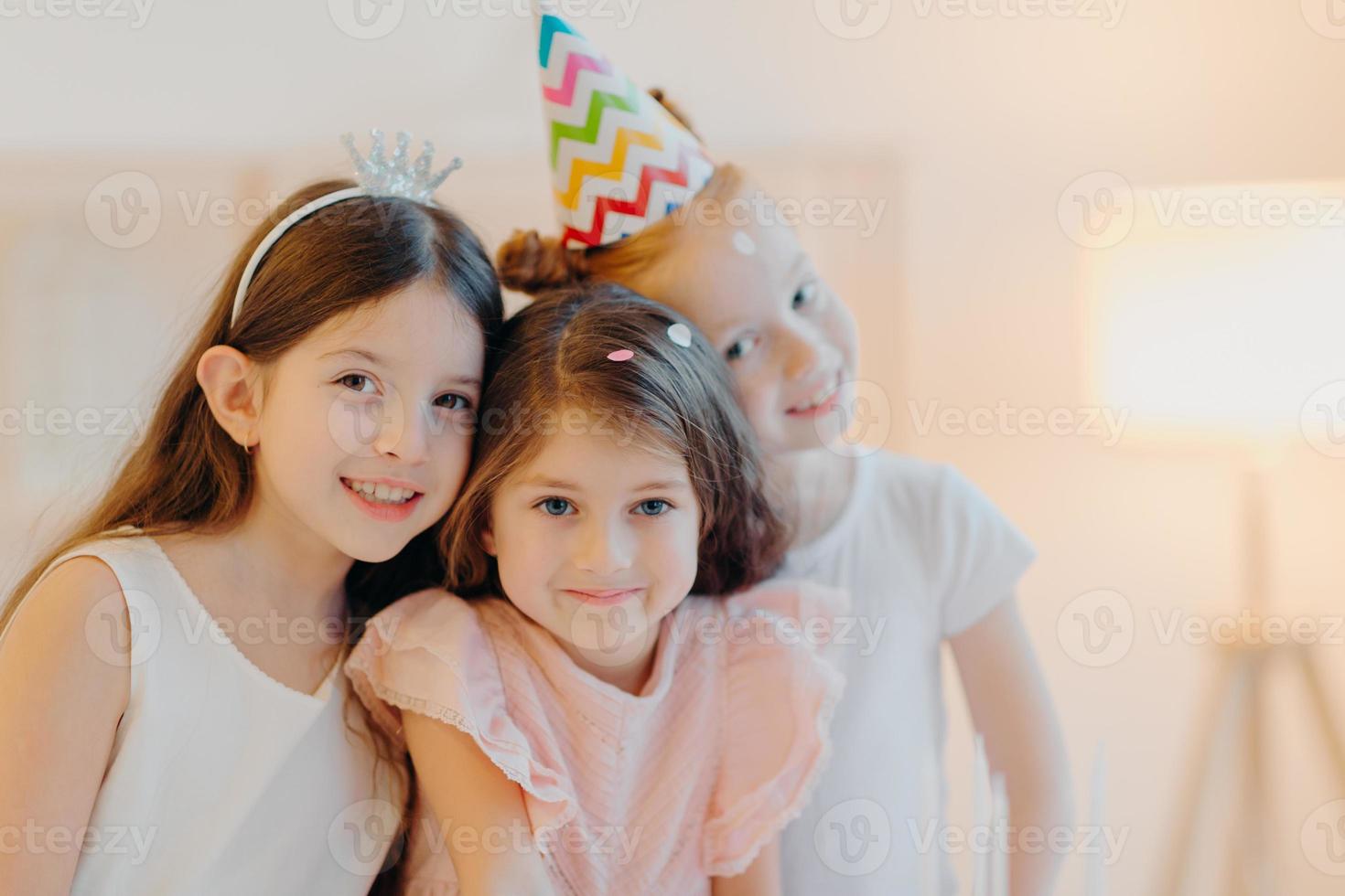 Portrait of happy three friends wear festive clothes, party hat, pose indoor against white background, play together during birthday celebration. Cute girls have good mood, come on special occasion photo