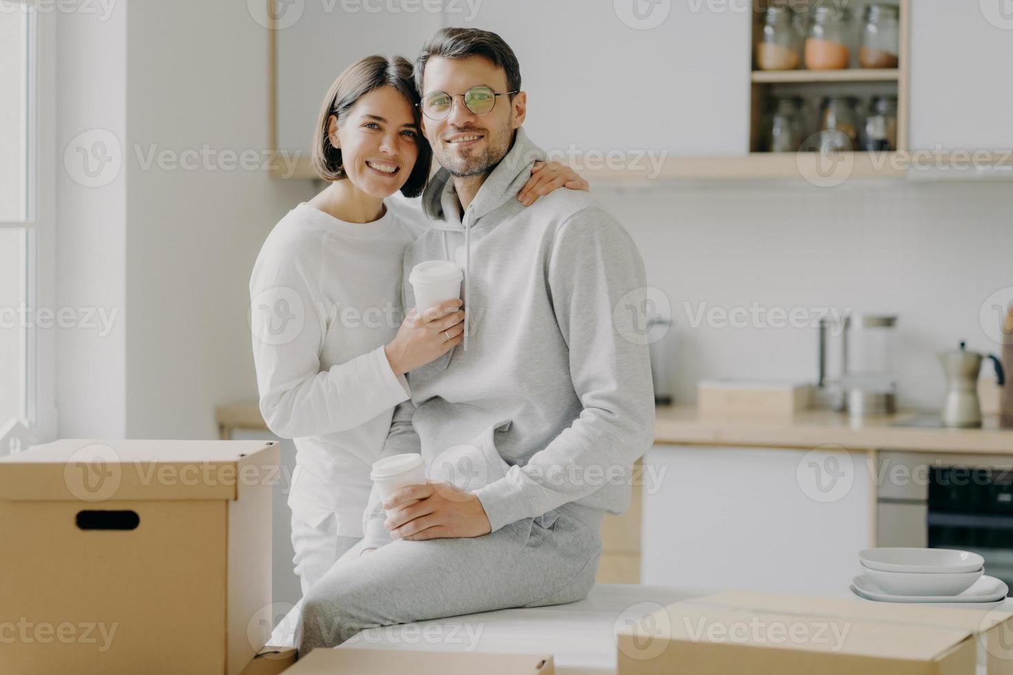 Image of glad family couple hug and stand closely to each other, drink takeout coffee, look with smile at camera, dressed in casual clothes, surrounded with cardboard boxes, spend free time in kitchen photo