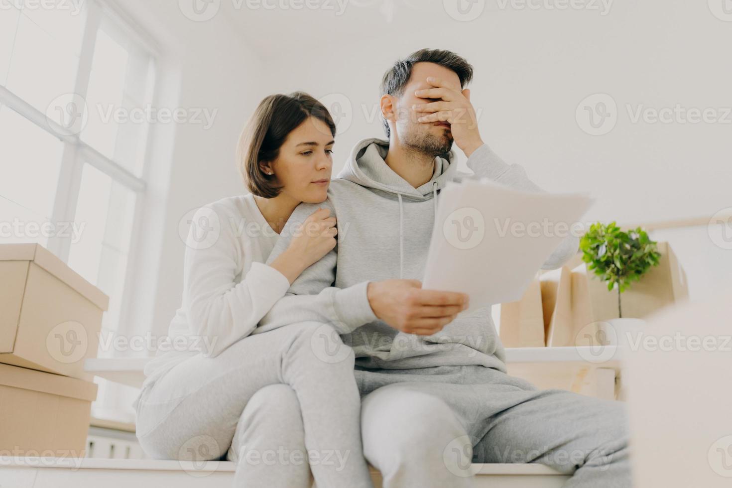 Dejected husband and wife manage finances, receive bill, face financial problem, have gloomy expressions, sit together in empty room, big window behind, cardboard boxes with personal stuff near photo