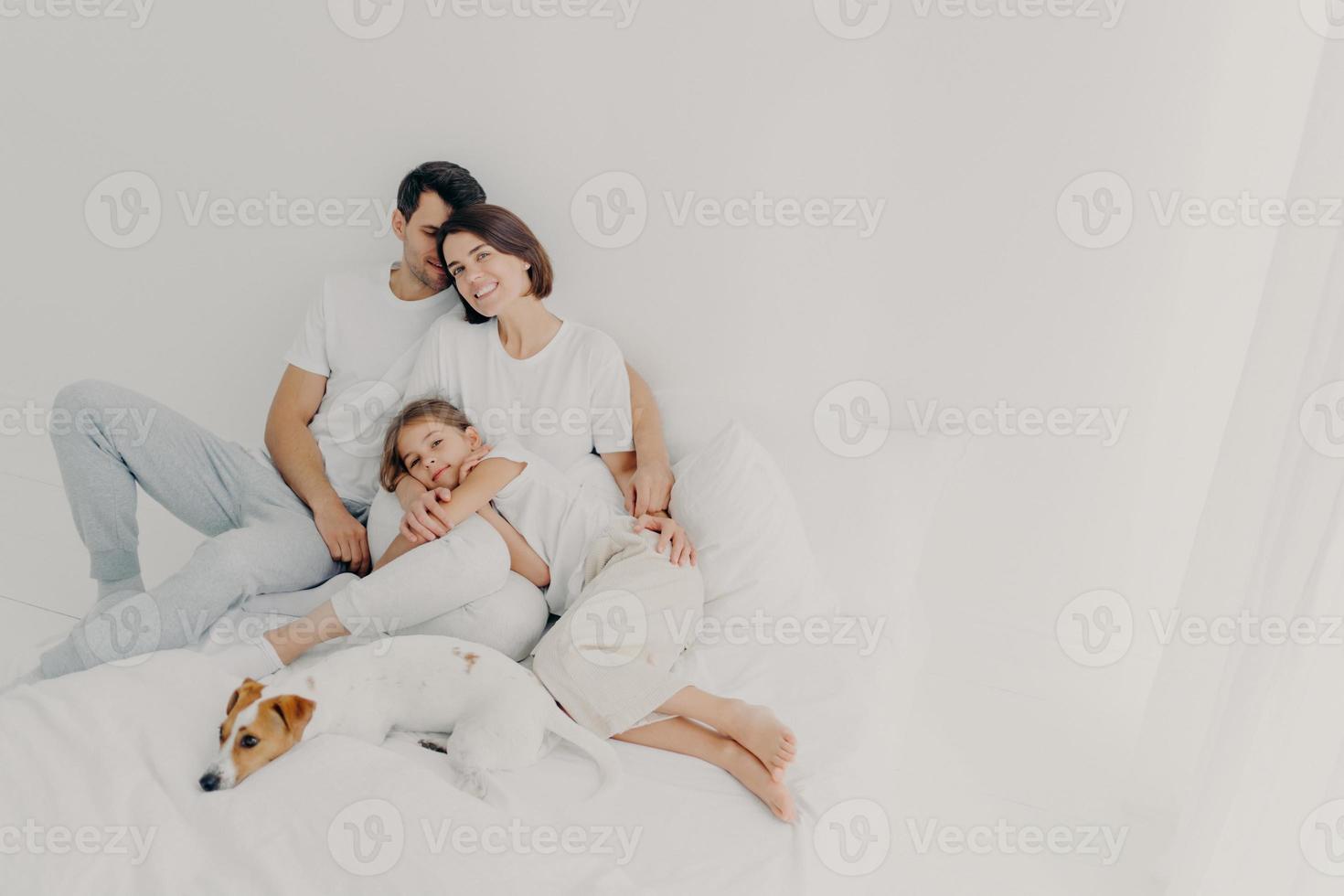 Pleased husband, wife and their small daughter pose in spacious bedroom with favourite pet, have fun, embrace and express love to each other, suppror in difficult situation. Family time concept photo