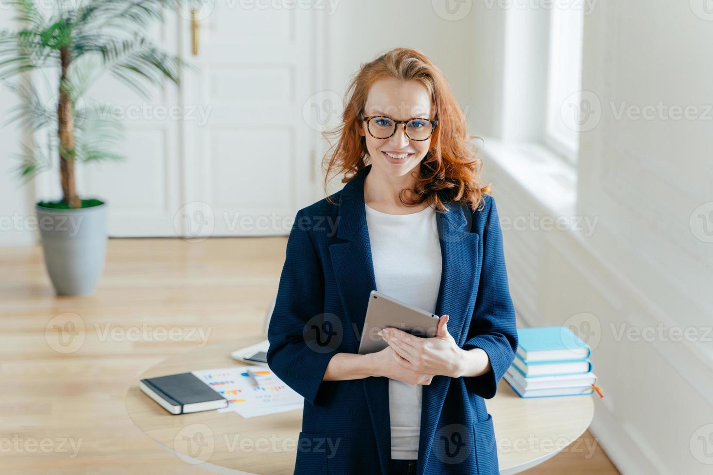 Horizontal shot of proud CEO checks email on digital touchpad, looks at camera with satisfied expression, wears spectacles, stands in modern office, enjoys online communication on modern device photo