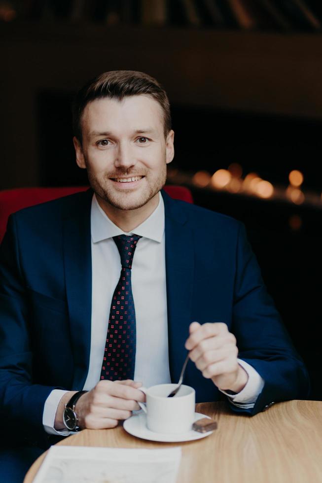 Satisfied male employee in luxury suit, drinks coffee, sits at wooden table in cafeteria, has pleasant smile and attractive look, waits for business partner or colleague going to discuss main problems photo