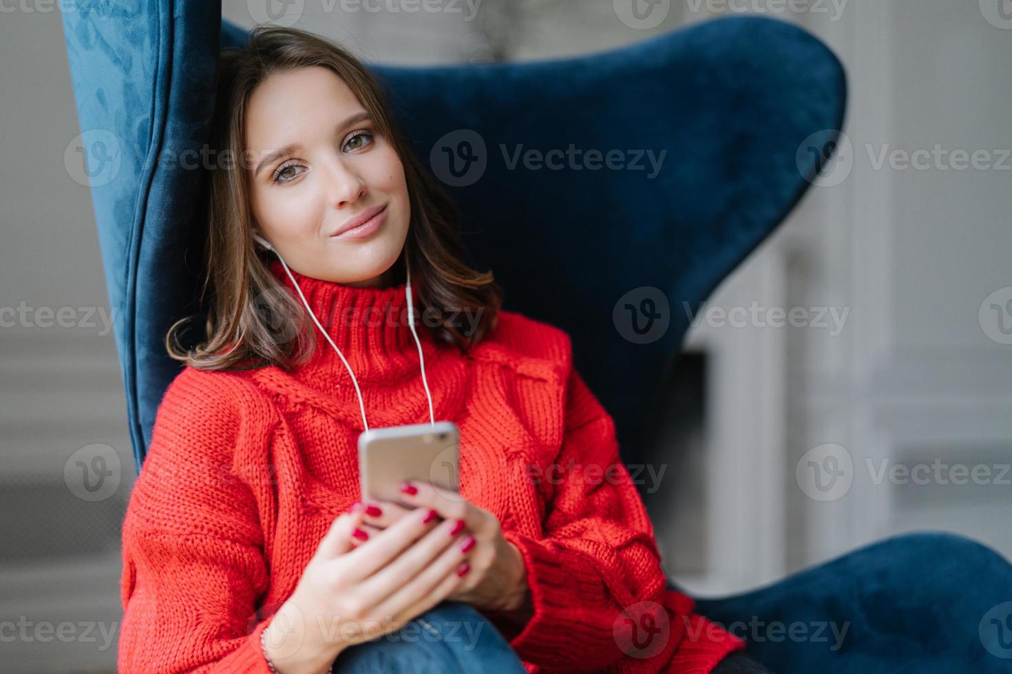 Pretty female teenager enjoys free time and popular music in earphones, holds modern smart phone, dressed in red knitted sweater, sits in comfortable armchair, uses application, checks new updates photo