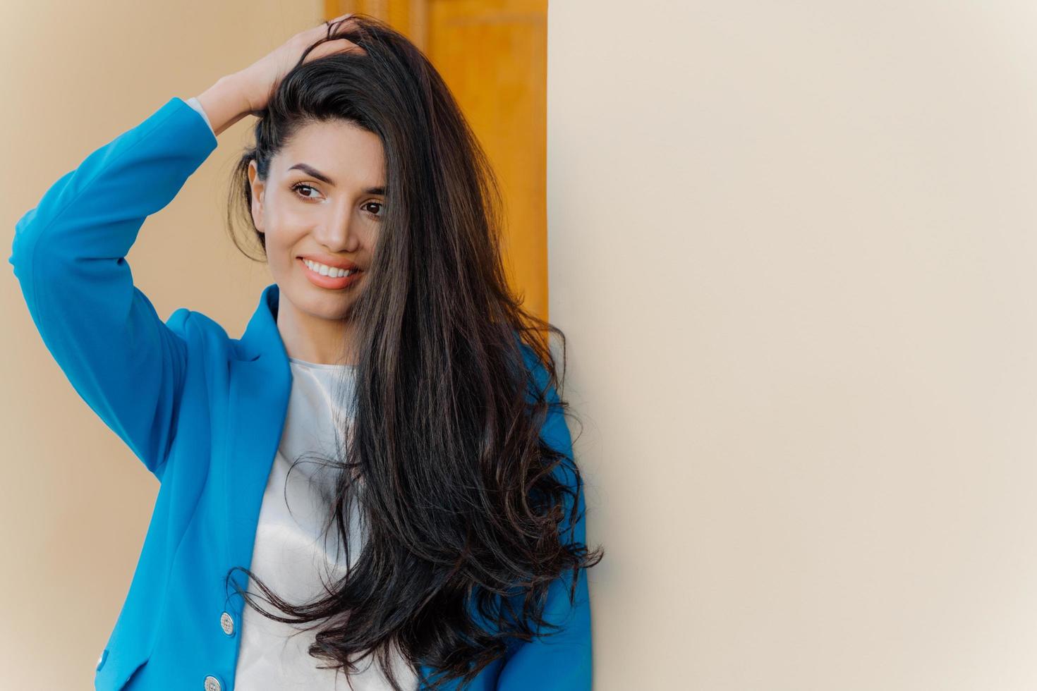 mujer morena encantadora con una sonrisa dentuda, mantiene la mano en la cabeza, mira a un lado, tiene maquillaje, se viste con un traje azul, se para cerca de una pared blanca, espacio en blanco para su contenido publicitario. dama elegante foto