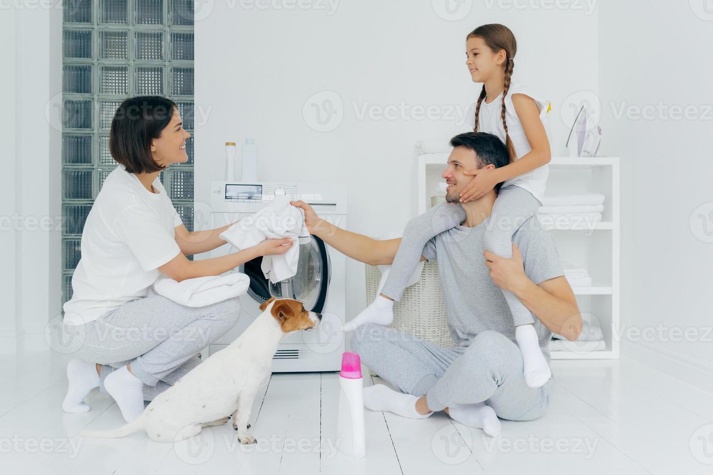 Family, housekeeping, washing concept. Smiling father gives piggyback ride to small daughter gives wife white t shirt to wash sits on floor at washing room in lotus pose, domestic animal near washer photo