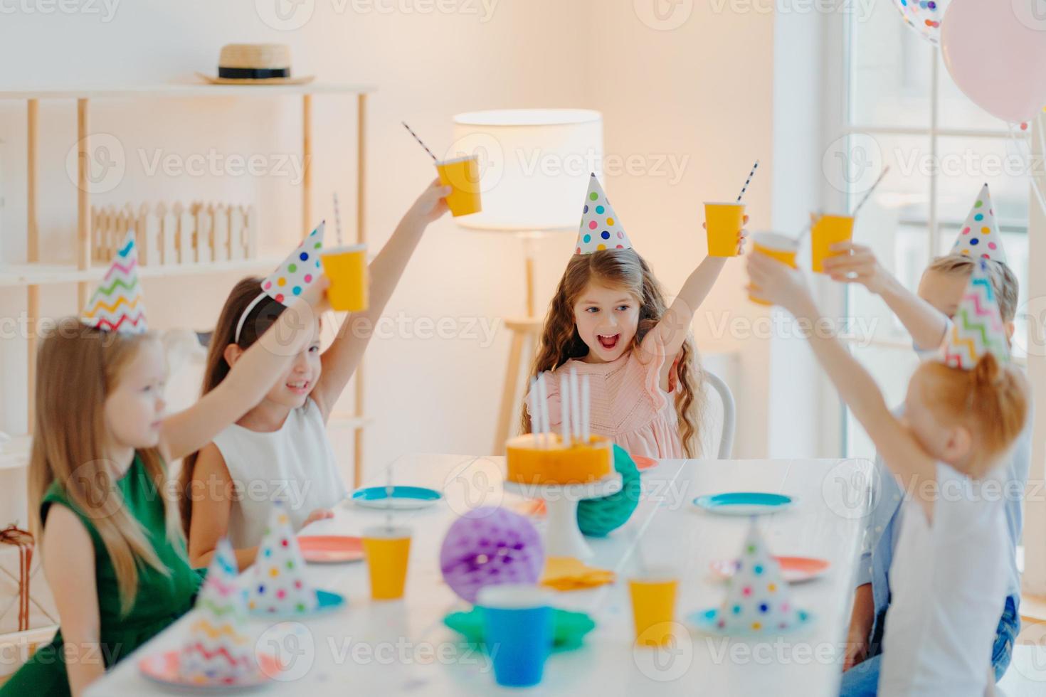 Group of cheerful preschool kids celebrate birthday together, have fun, cheer with cups of beverage, wears festive hats, eat delicious cake, sit at table in spacious room. Children and party concept photo