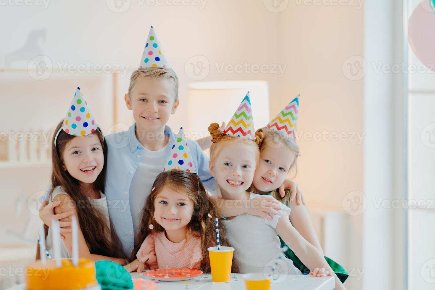 Group of adorable kids wear party hats, embrace and have fun, celebrate birthday, pose in decorated room, gather near festive table, have fun cuddle and look gladfully at camera photo