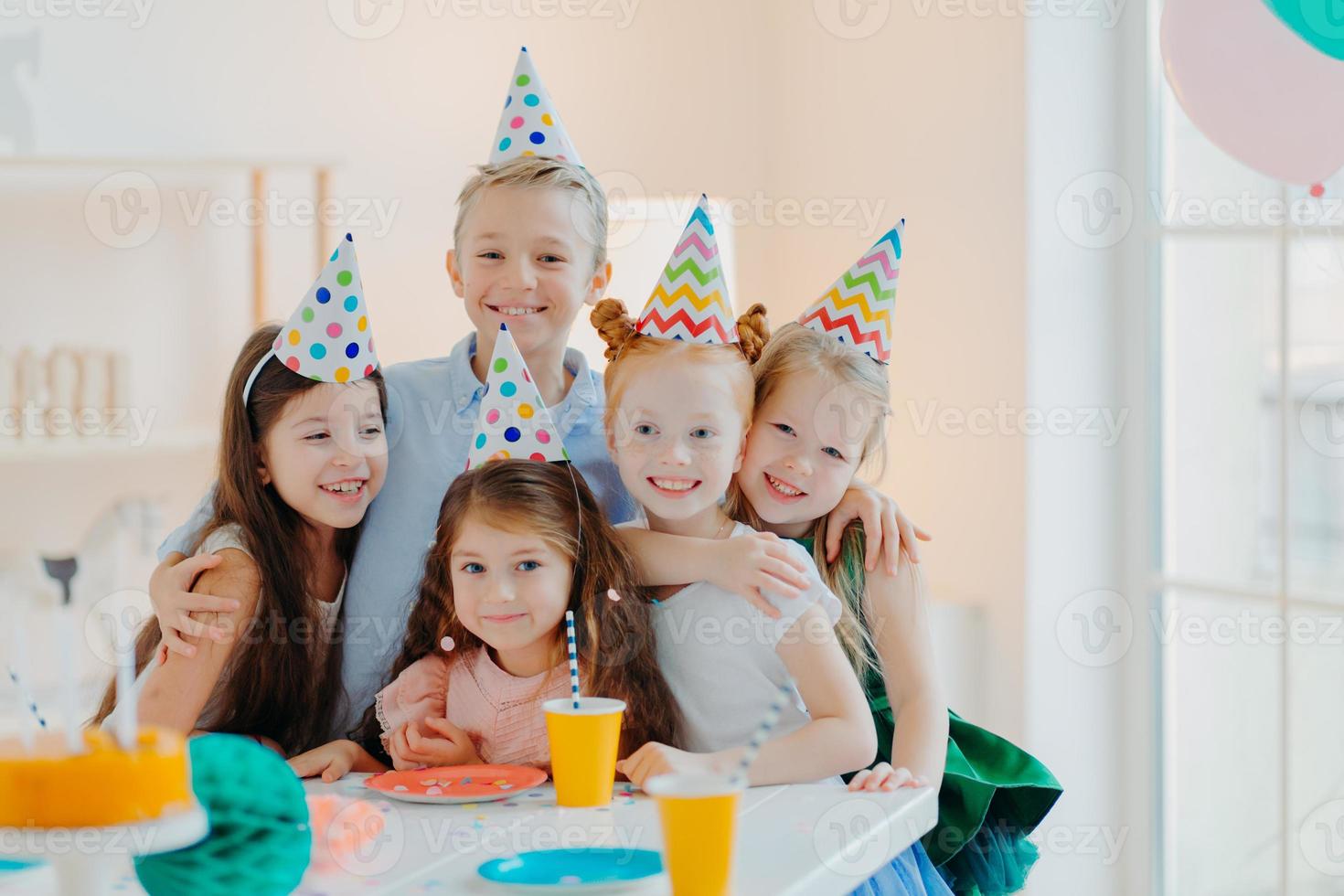 Five friendly little children wear festive cone caps, hug and make photo together, play games and celebrate birthday, have glad expressions, pose at festive table