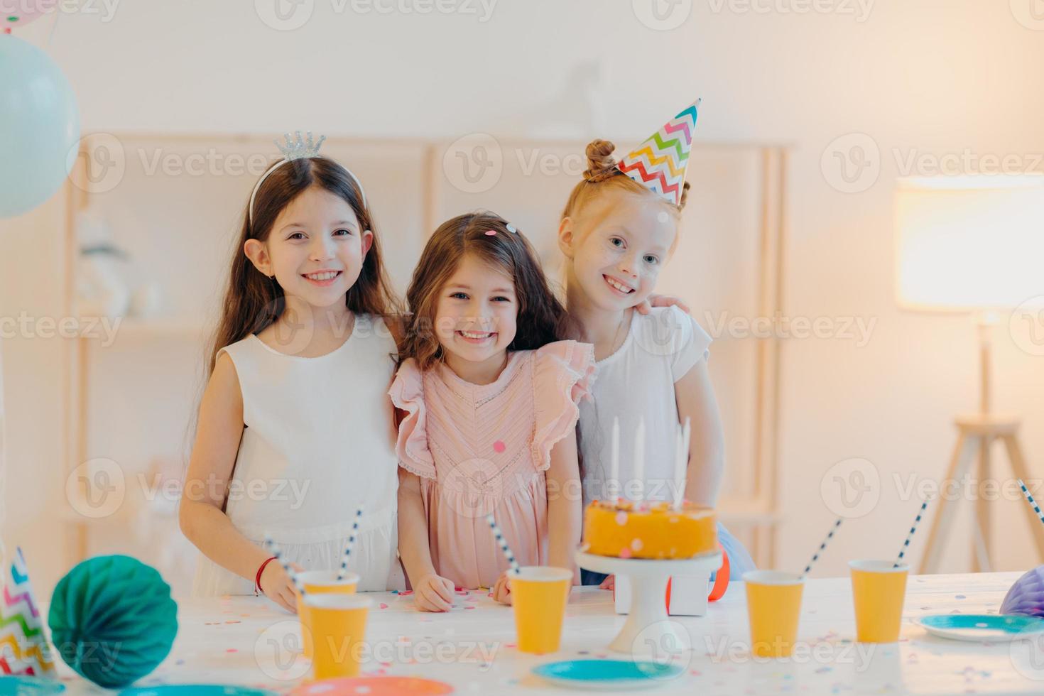 Indoor shot of happy three girls embrace and have fun, smile gladfully, stand near festive table with cake, cups, have happy childhood, being on party together. Childhood and festivity concept photo