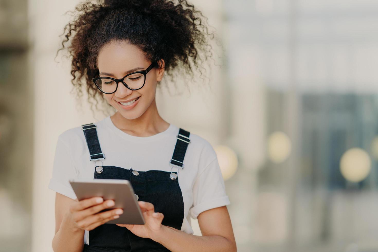 foto de una atractiva chica del milenio con el pelo terso, sostiene una tableta digital, navega por las redes sociales, usa anteojos ópticos, camiseta informal y sarafan, conectada a internet inalámbrico, escribe un mensaje