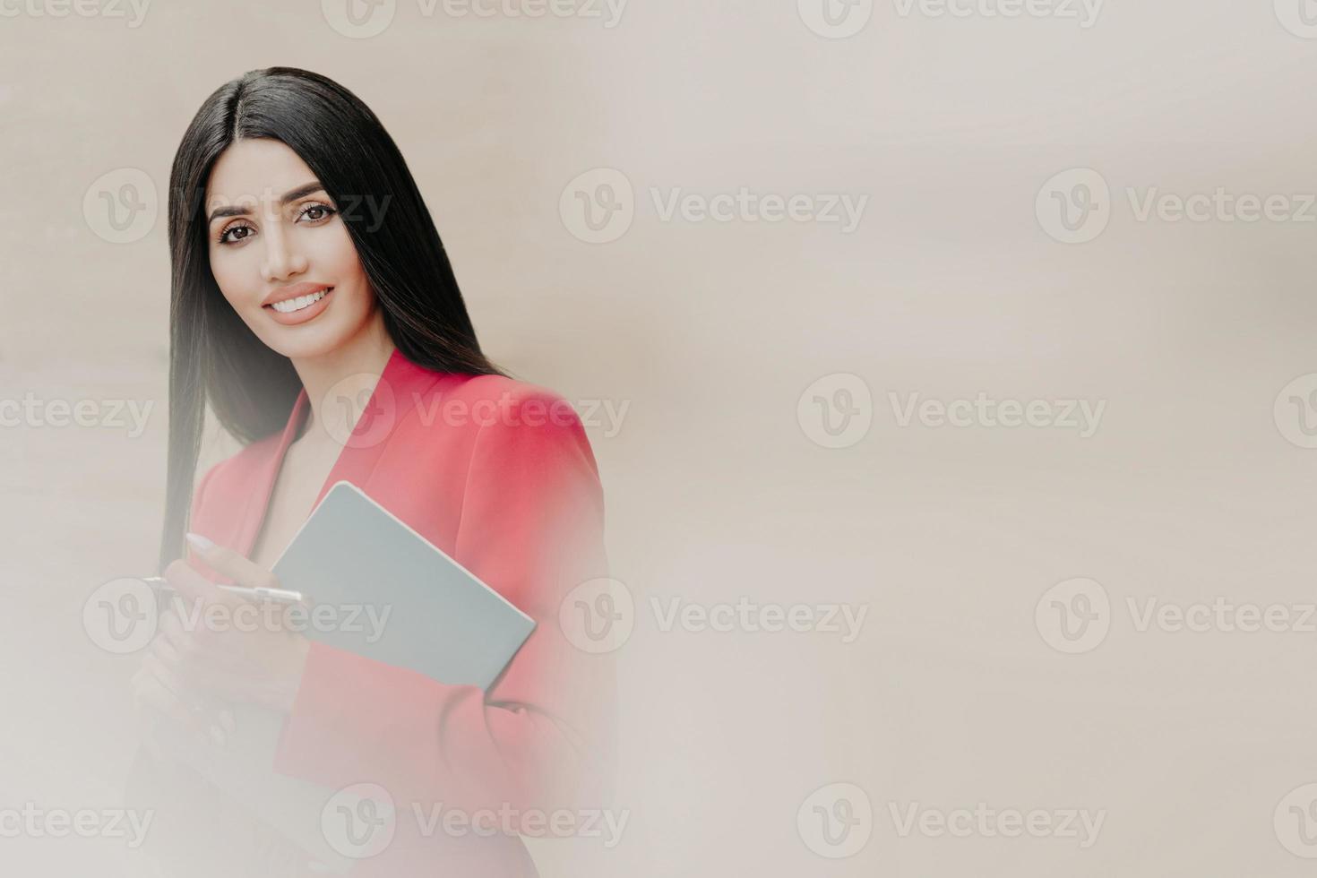 Horizontal shot of lovely brunette woman with happy facial expression, dressed in formal clothes, holds notebook and pen, has black straight hair, stands over white blurred background with free space photo