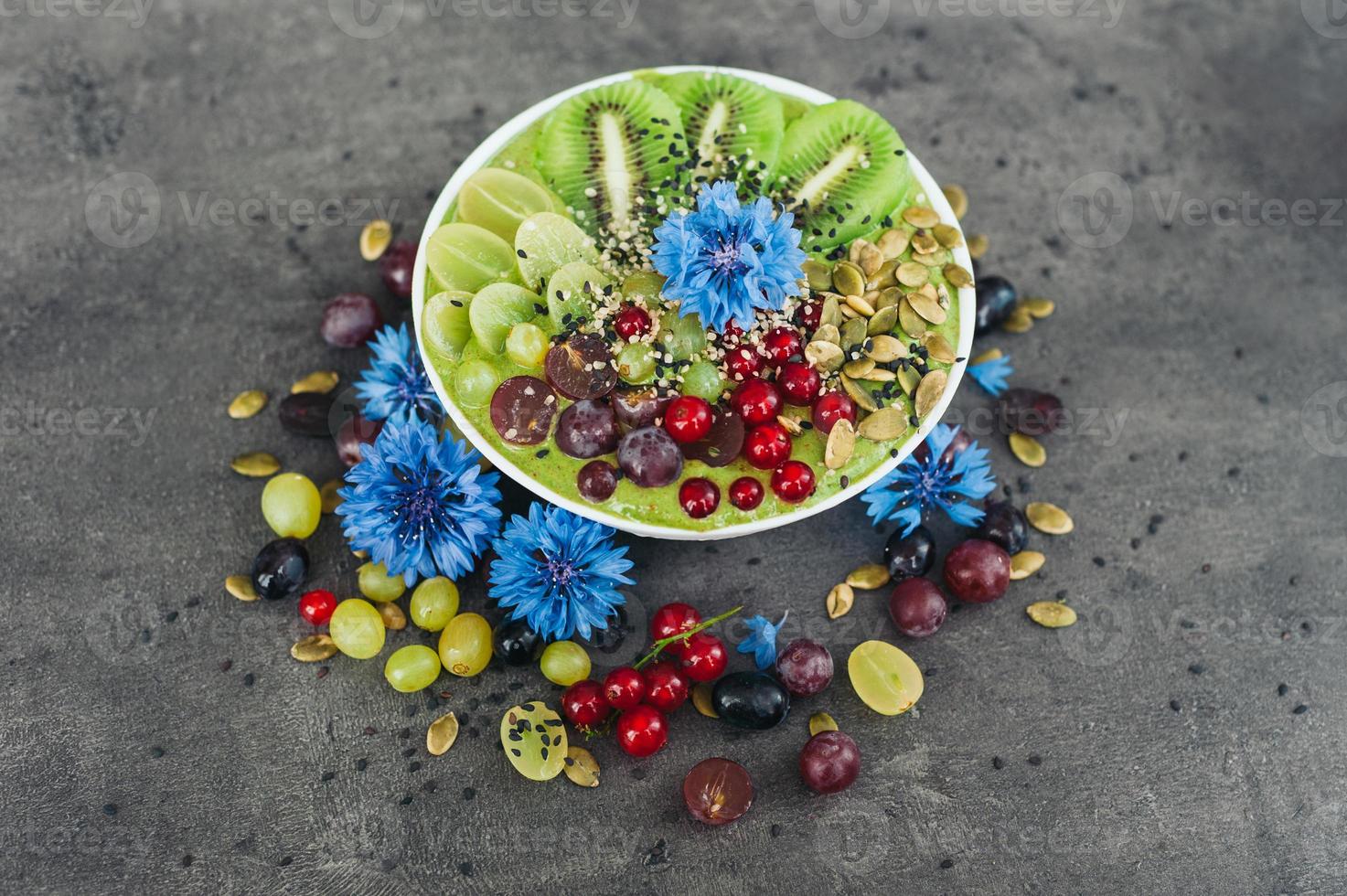 Bowl with fresh kiwi salad, seeds, red current, green grape and blue cornflowers around. Vegeterian dish of ripe fruit. Top view. Blank space photo