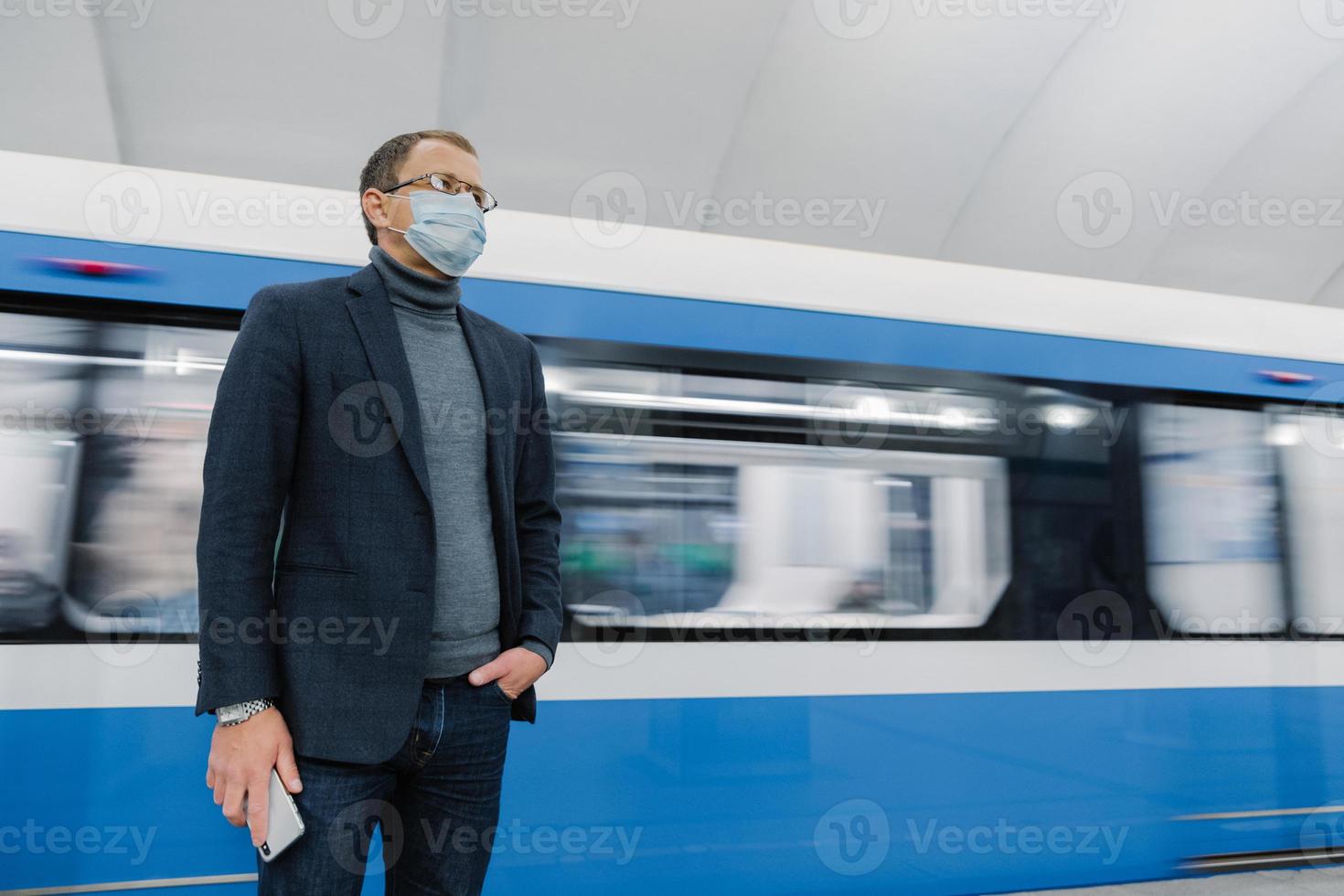Pensive businessman wears surgical sterile mask to prevent spread of respiratory disease, poses against underground train, travels in public transport, holds smartphone in hand. Coronavirus outbreak. photo