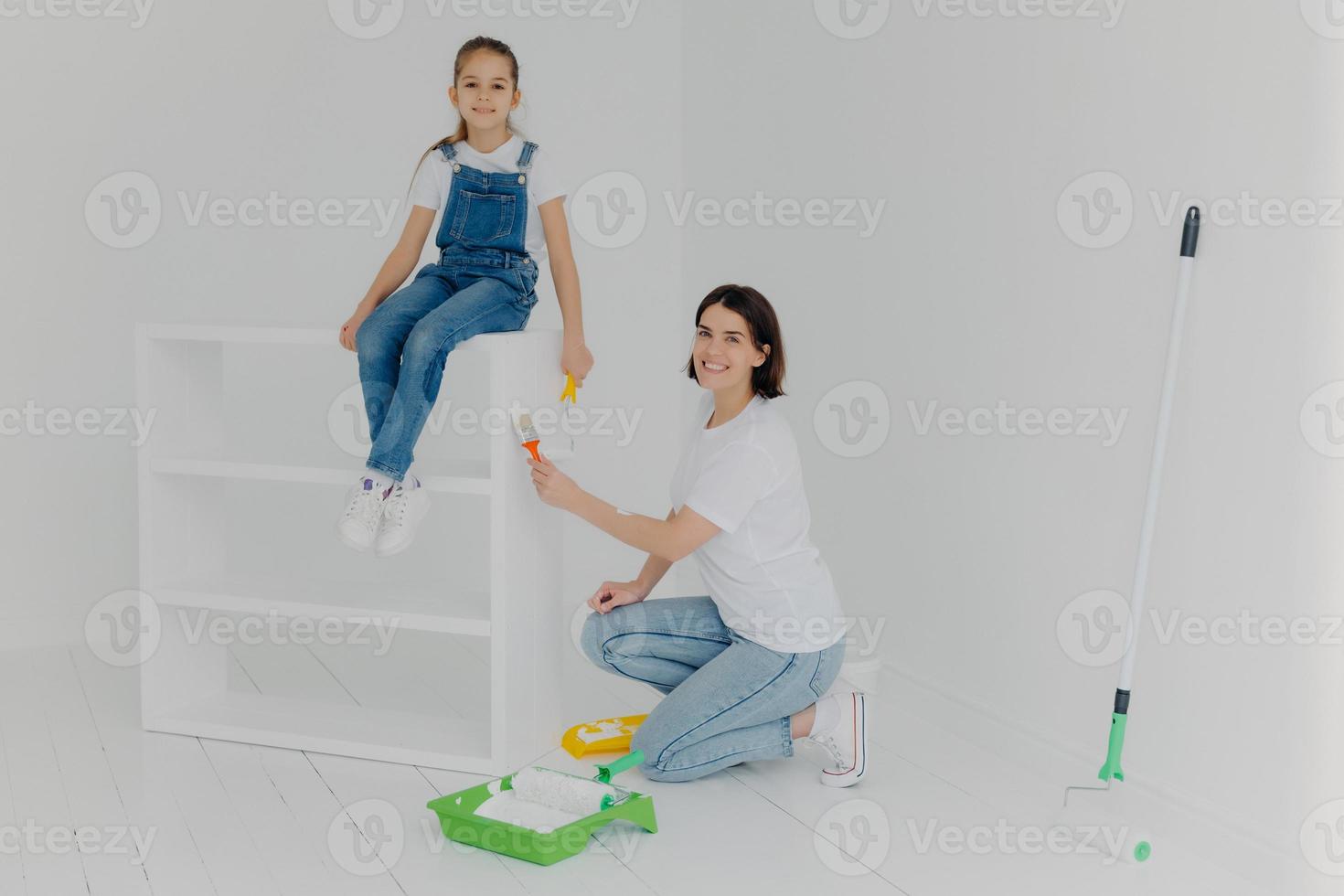 Shot of lovely mother and small hard working daughter pose in empty room, paint furniture in white color, use paintbrushes, happy to finish work, being busy during weekend. People, repair, improvement photo