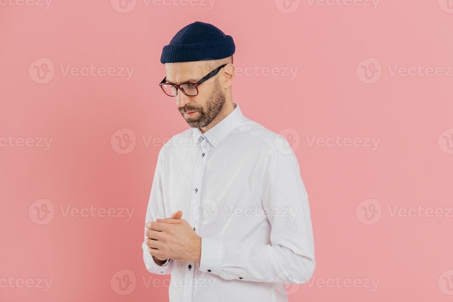 Contemplative unshaven man keeps gaze down, wears white shirt, hat and glasses, being deep in thoughts, tries to make solution, thinks how develop his business, isolated over pink background photo