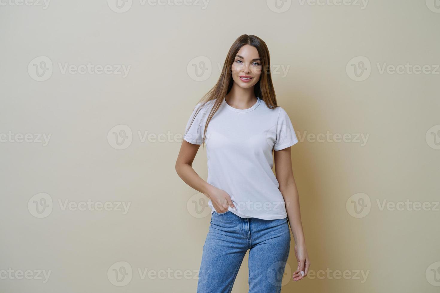 Studio shot of pretty slim woman keeps hand in pocket, wears casual t shirt and jeans, stands in relaxed pose, looks at camera self confident, isolated over beige background talks casually with client photo