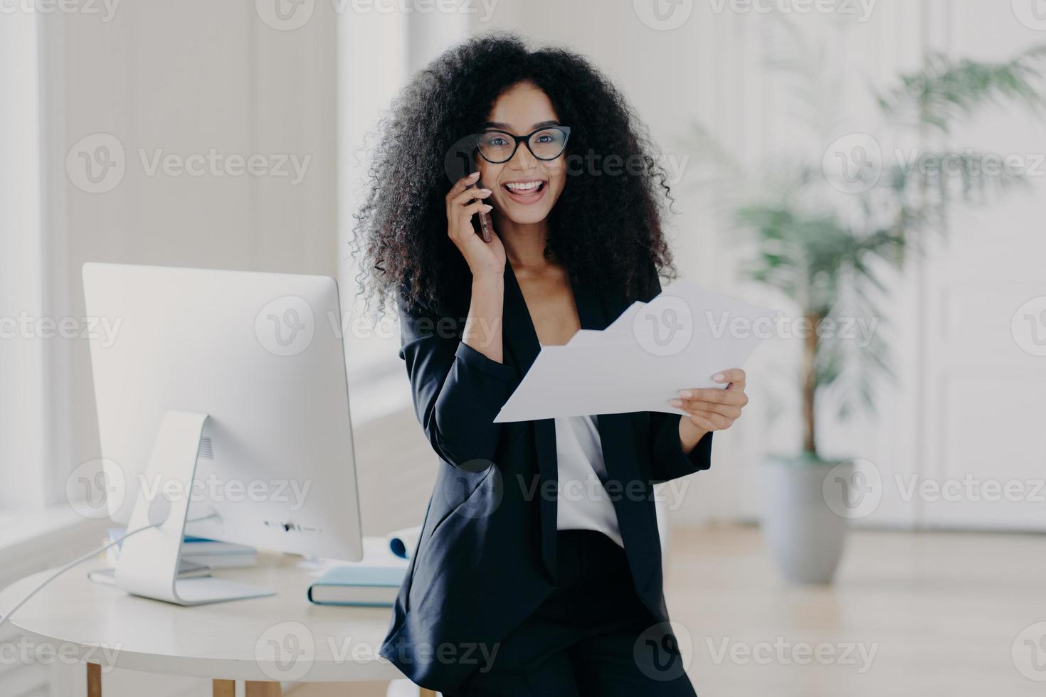 Intelligent woman with Afro hairstyle, holds paper documents, checks information, calls business partner via cell phone, analyzes future strategy, speaks with banking manager, dressed in black suit photo