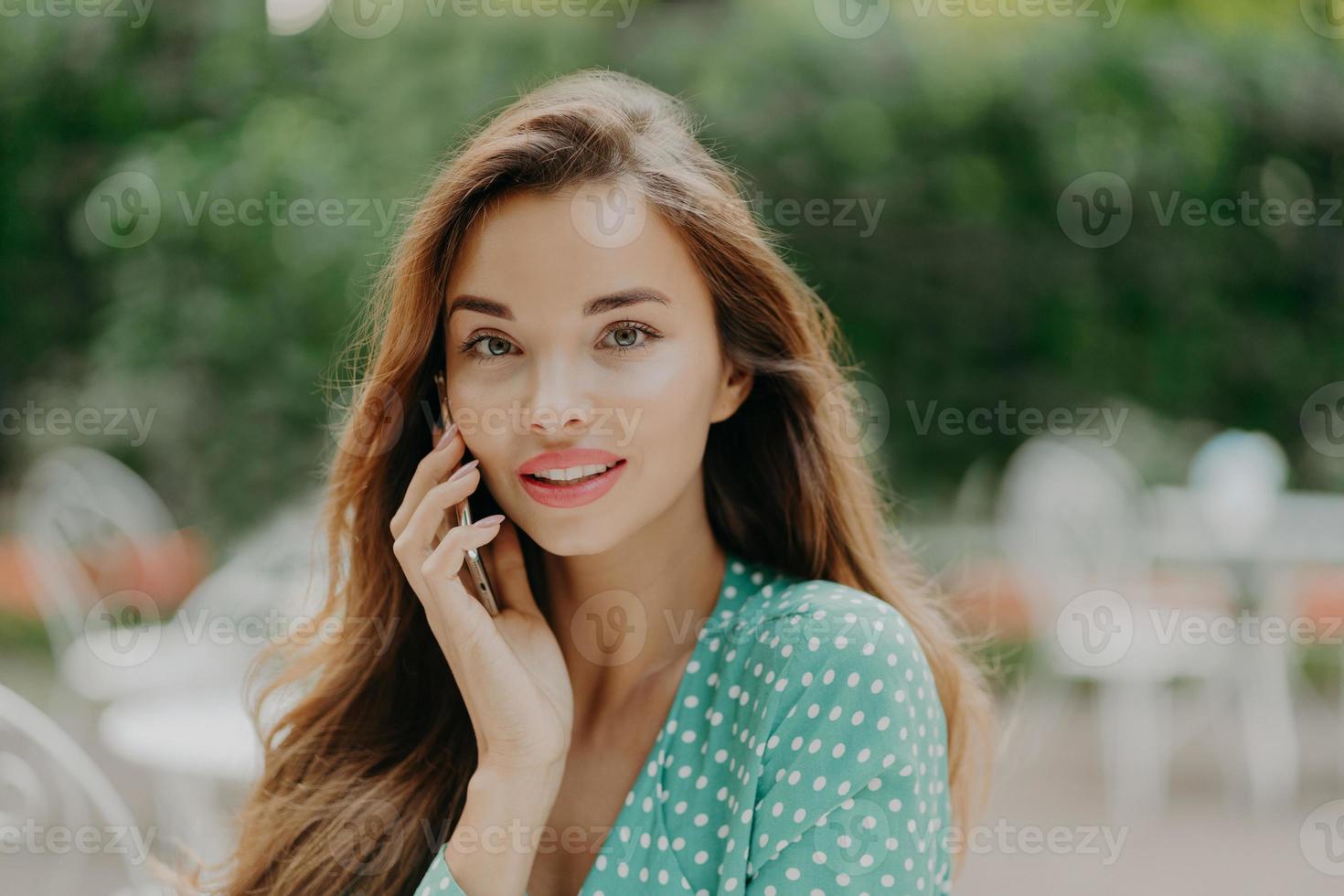 Horizontal shot of pretty young woman wears polkadot blouse, makes phone call, speaks with friend, poses outside against green blurred background, wears lipstick. People and communication concept photo