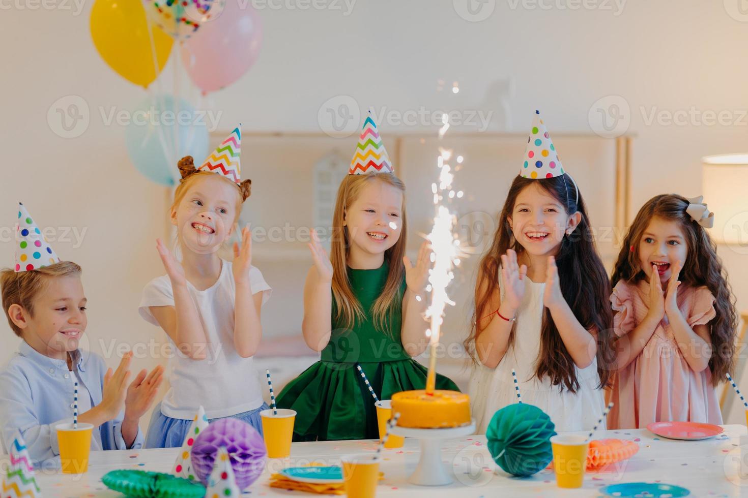 concepto de vacaciones y eventos festivos. felices cinco niños pequeños aplauden, miran el brillo del pastel, celebran el cumpleaños de sus amigos, se reúnen cerca de una mesa grande, se paran en una habitación decorada con globos foto