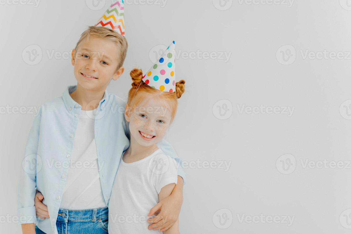 Happy small ginger freckled girl and little boy embrace and smile gladfully, wear party hats, enjoy nice time together, isolated over white background, copy space. Children and festive event concept photo