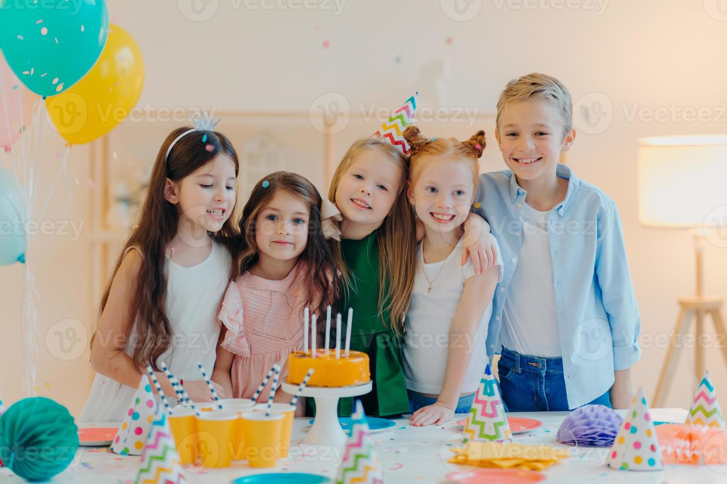 Horizontal shot of group of little children gather together to celebrate birthday, embrace and pose at camera, prepare for special occasion, stand near table with cake, paper cups, party caps photo