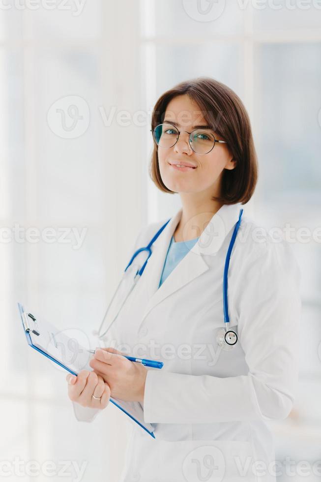 Qualified cardiac expert with stethoscope prepares for health seminar, gives consultancy about first aid, writes down information in clipboard, wears white gown, ready to give medic care help photo