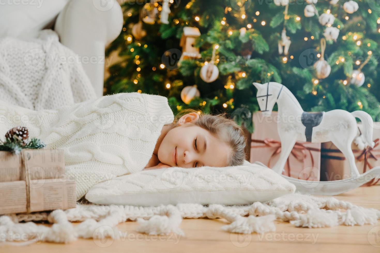 Charming girl sleeps on soft white pillow on floor against decorated New Year tree, has pleasant dreams, surrounded with toy horse and gift boxes. Children, rest and winter holidays concept. photo