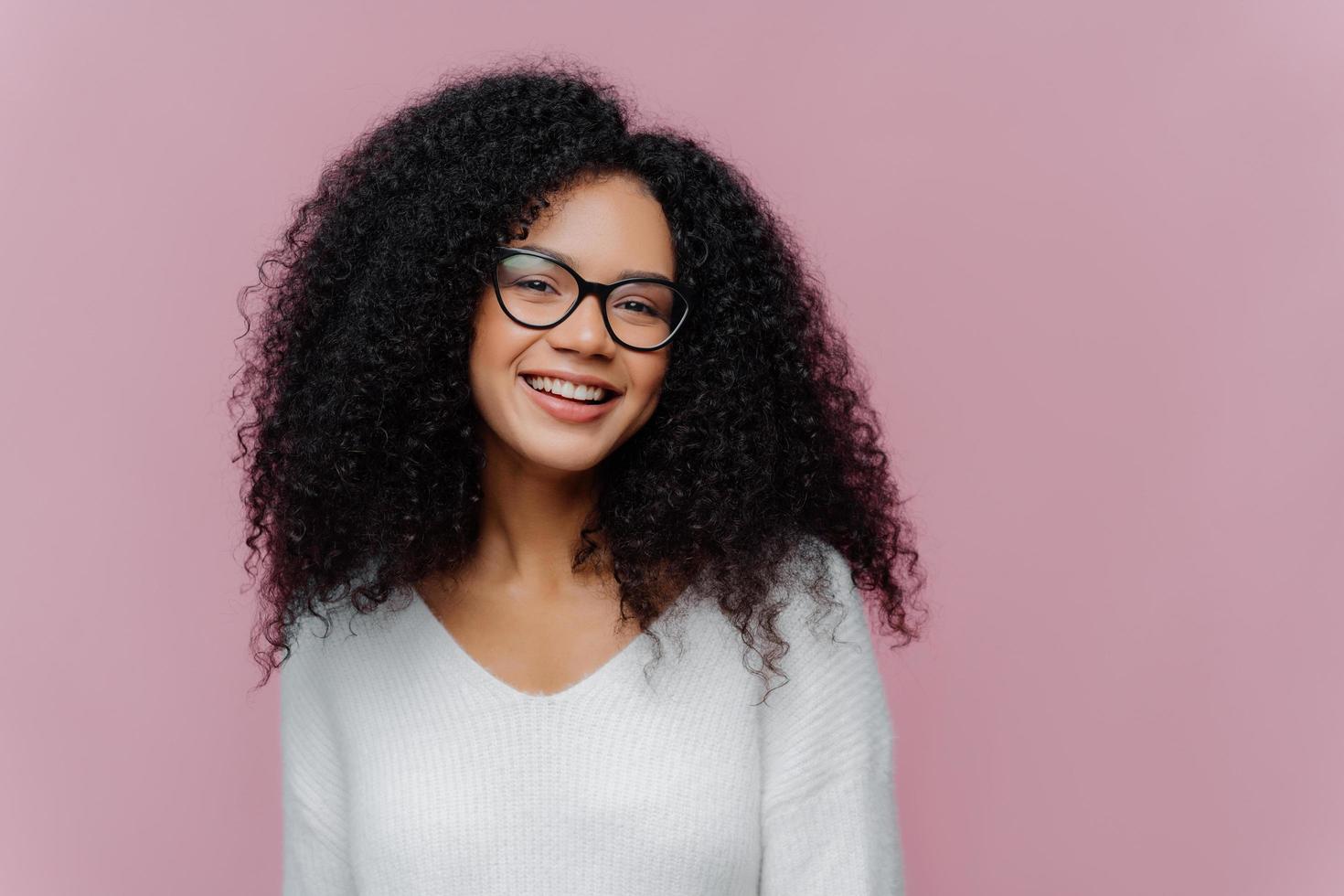 Headshot of pleased lovely woman with curly hairstyle, smiles gently at camera, wears optical glasses and white casual sweater, poses against purple background. Positive emotions and feelings concept photo