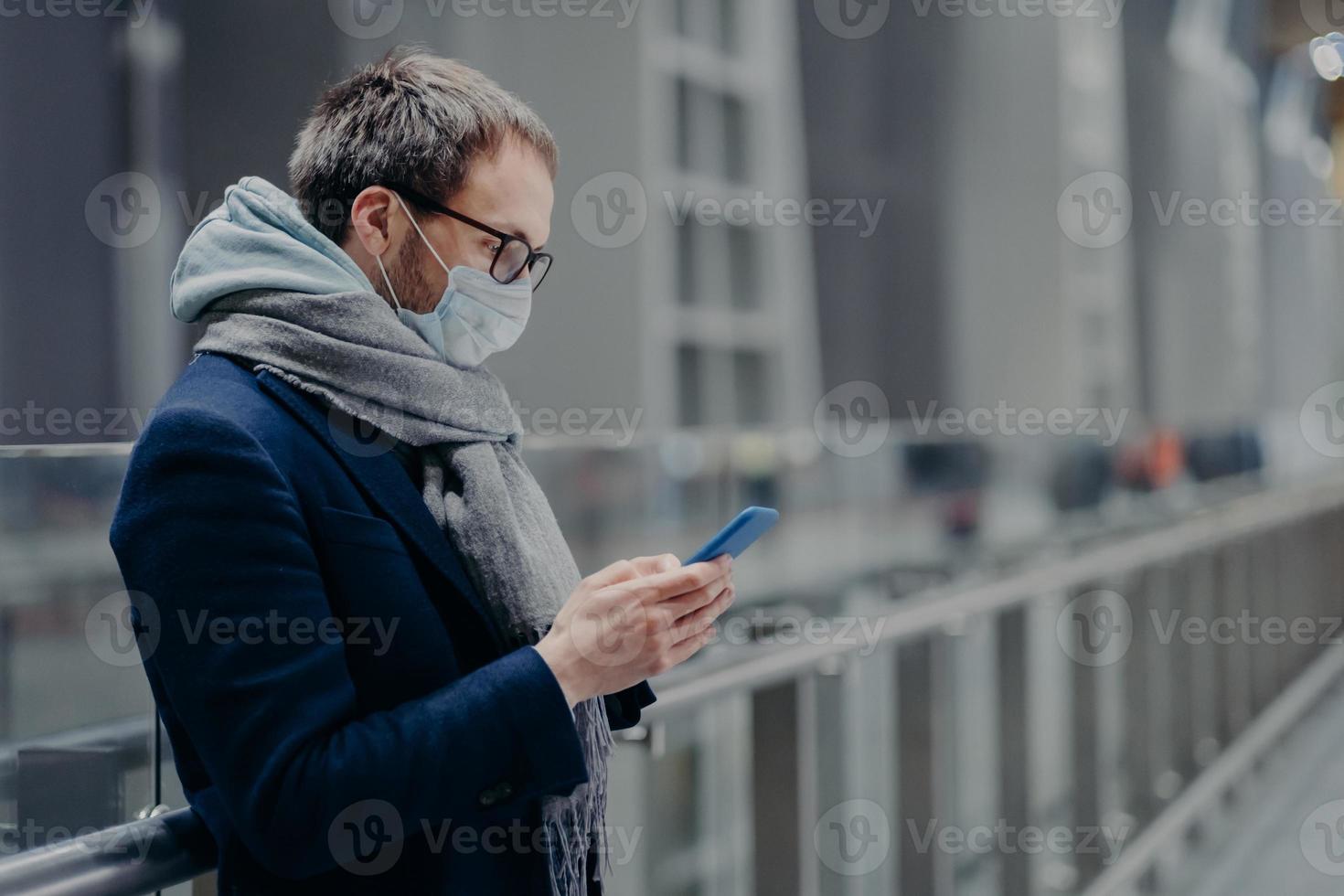 Sideways shot of male blogger sends text messages and reads comments from followers on modern cellphone, wears sterile medical mask to protect himself from coronavirus or other infectious disease photo