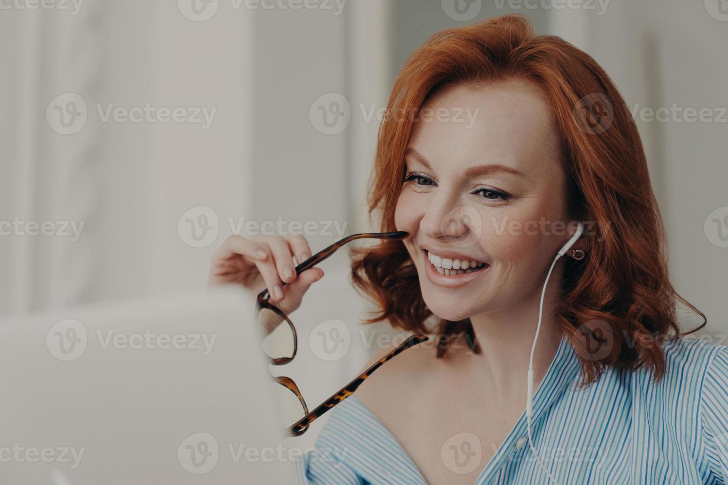 Beautiful cheerful redhead woman uses laptop computer for online conference, talks with colleagues distantly, holds spectacles, discusses working issues, works freelance from home. Technology concept photo