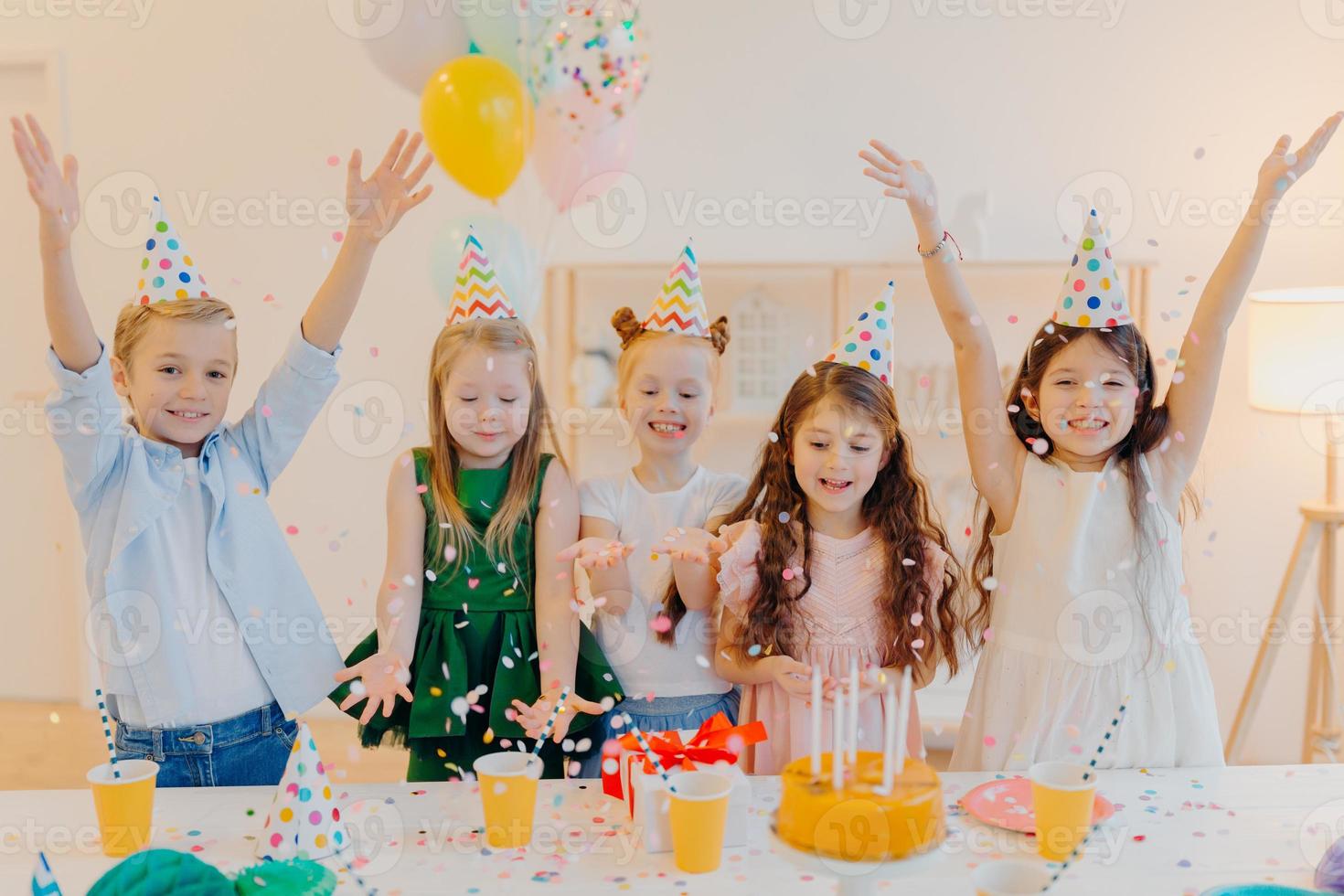 Horizontal shot of happy positive children catch confetti, celebrate birthday together, raise arms, have good mood, play together, stand near festive table with present box, cake, party hats photo
