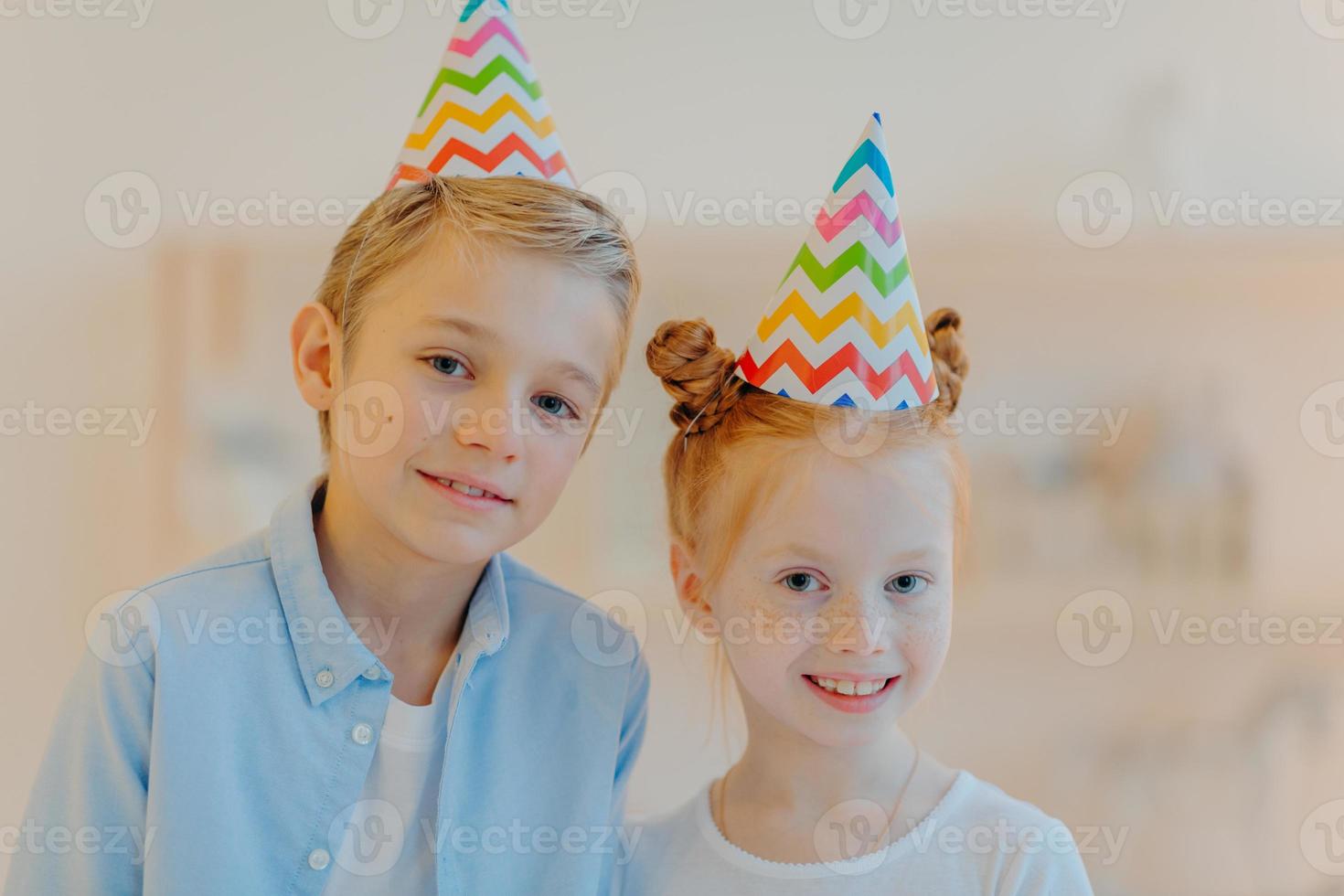 Portrait of happy ginger girl and her broghter stand closely to each other, wear party hats, come on friends birthday, look gladfully at camera, pose against blurred background. Childhood concept photo