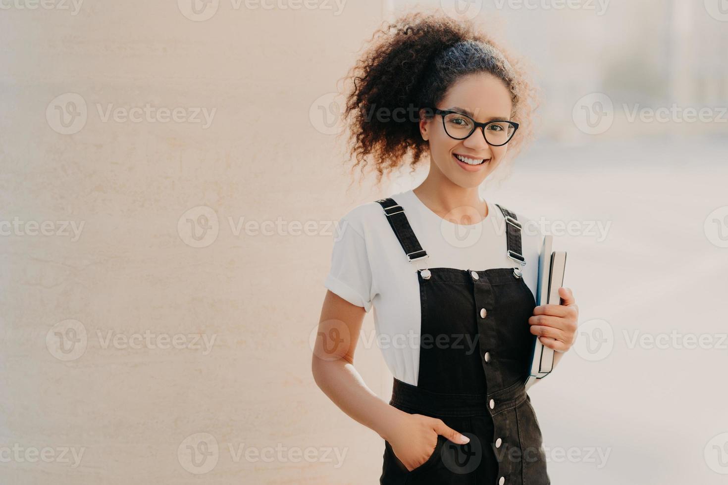Lovely curly woman with combed hair, dressed in white t shirt, sarafan, keeps hand in pocket, holds book and textbook, wears optical glasses, poses at unversity campus, smiles happily at camera photo