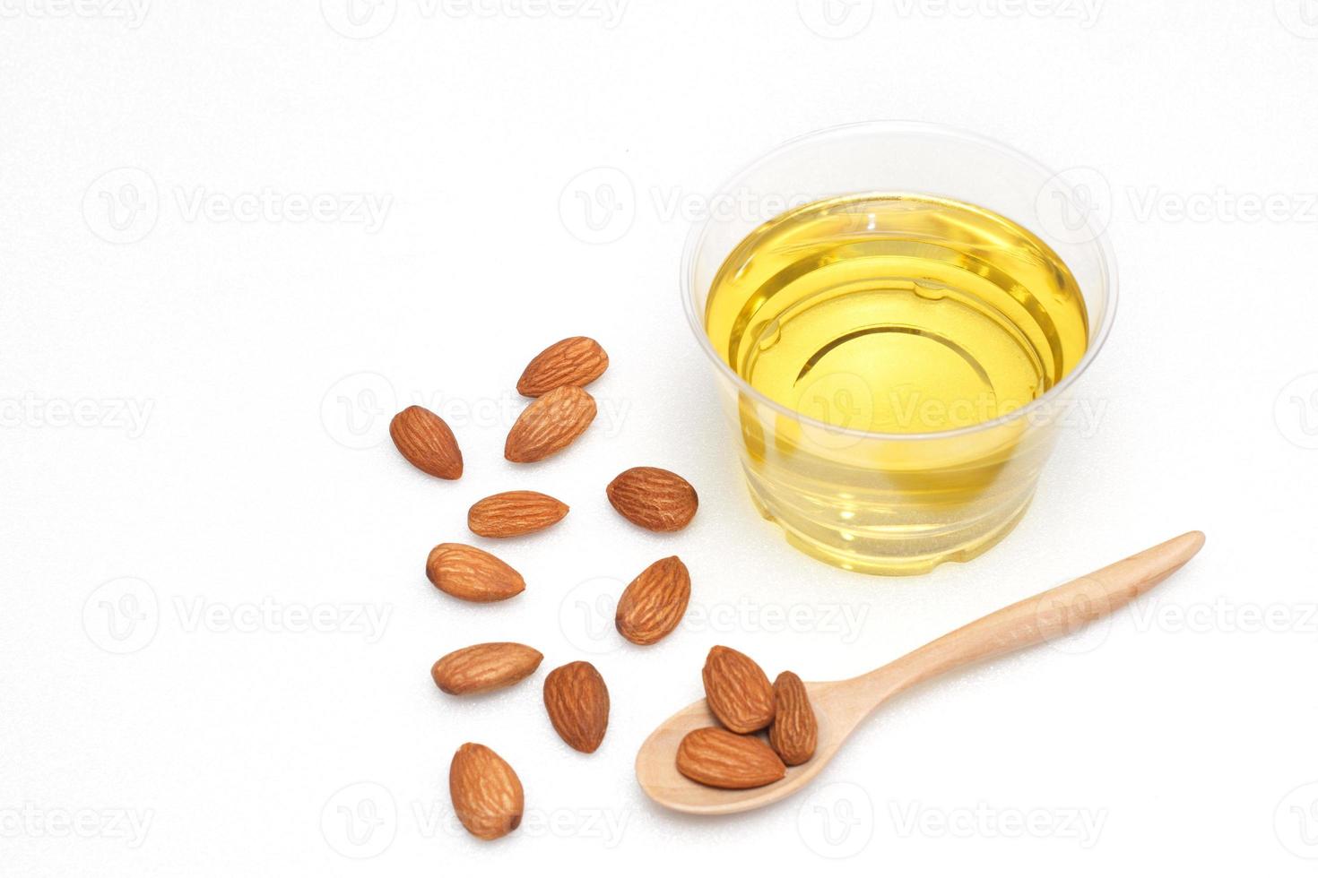 Yellow almond oil in a clear bowl with a wooden spoon next to it and several almonds on a white background photo