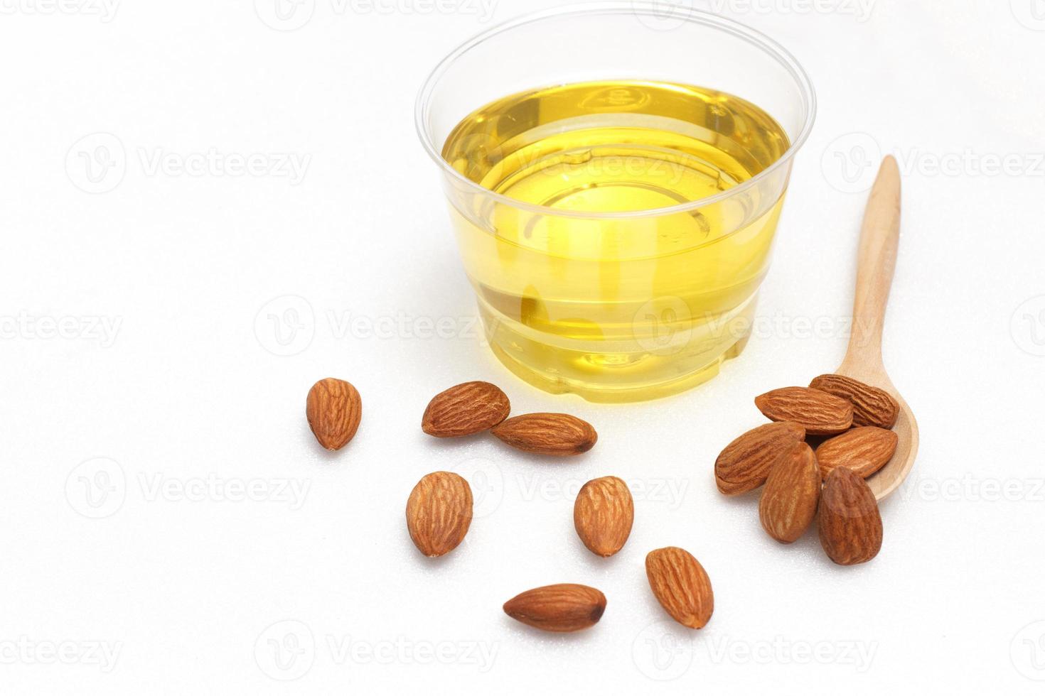 Yellow almond oil in a clear bowl with a wooden spoon next to it and several almonds on a white background. photo