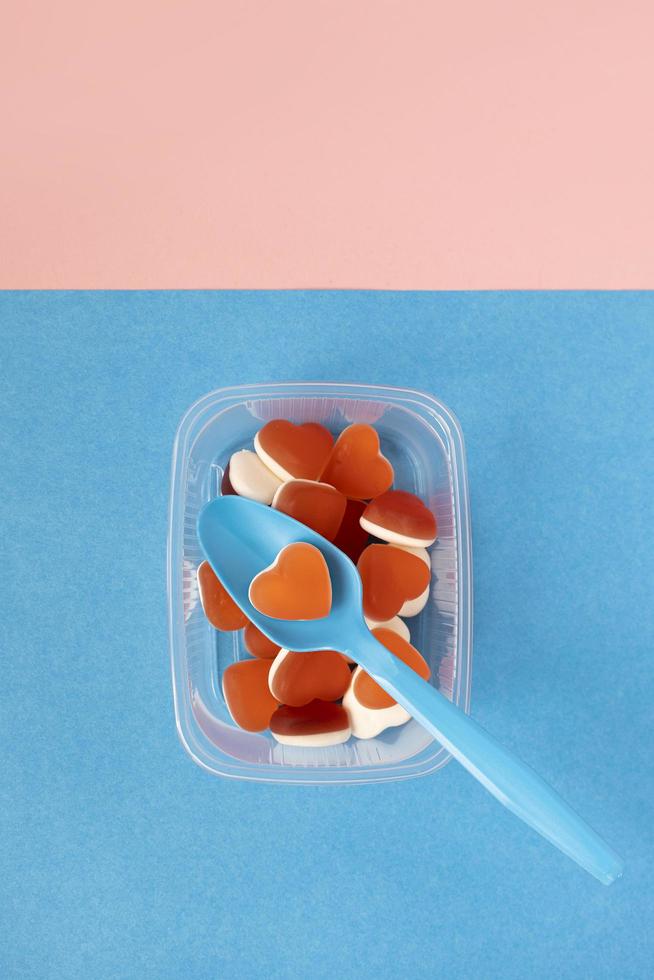 Sweet jelly candies and spoon in transparent plastic plate on blue background. Heart shaped marmalade. Concept of food, fast food, romantic lunch. Top view. photo