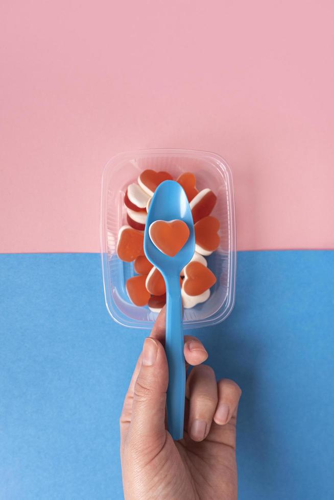 Female hand holds plastic blue spoon with red heart shaped jelly marmalade. Tasty candy in transparent plastic plate on blue and pink background. Concept fast food. Top view. photo