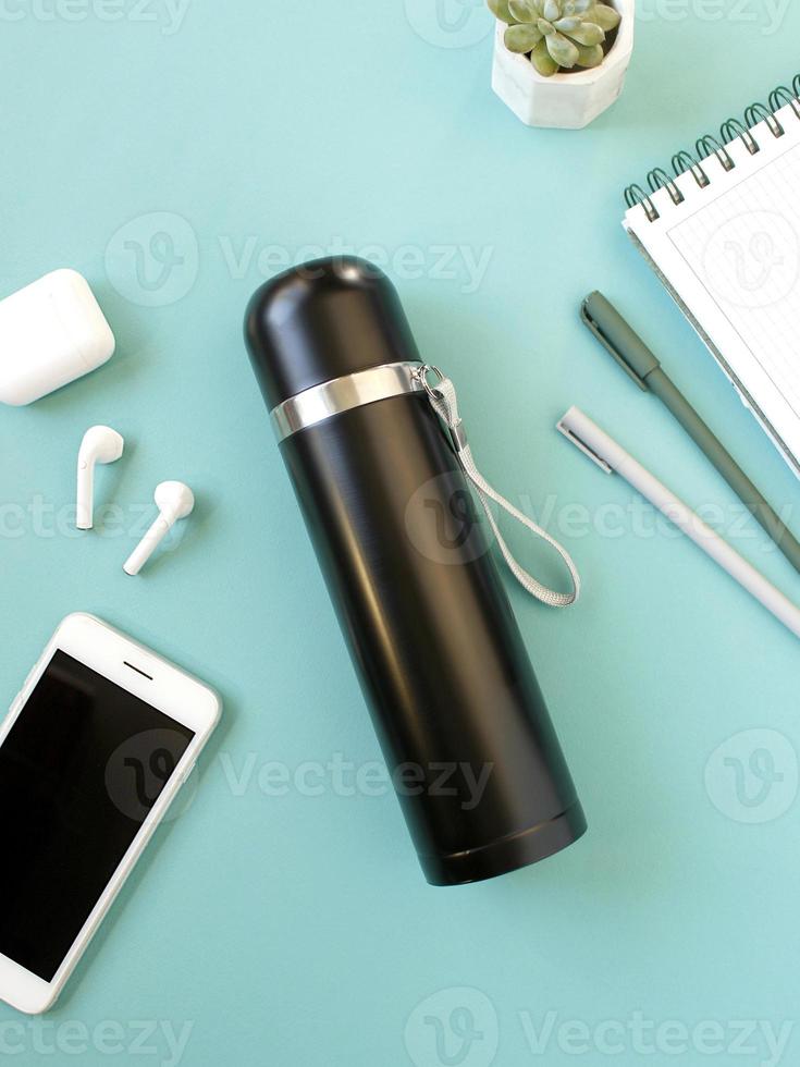 Freelancer workplace, stylish coffee drinks from thermos and takes notes, smartphone with earphones and ecologic steel thermo bottle for water. photo