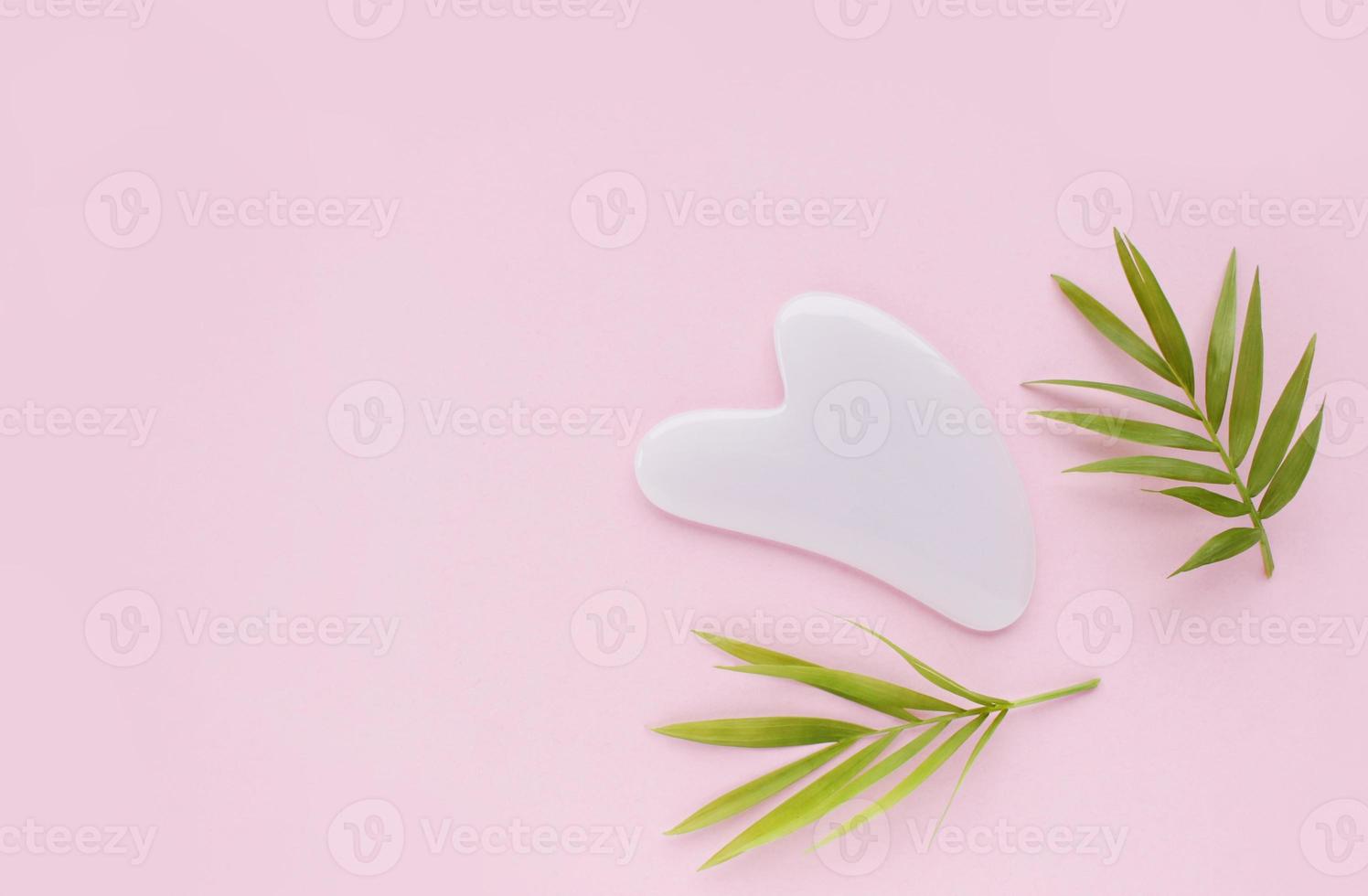 Top view of pink heart shaped guasha stone made of quartz crystal on pink background with copy space. Concept of alternative skin care treatment, self massage and acupressure photo