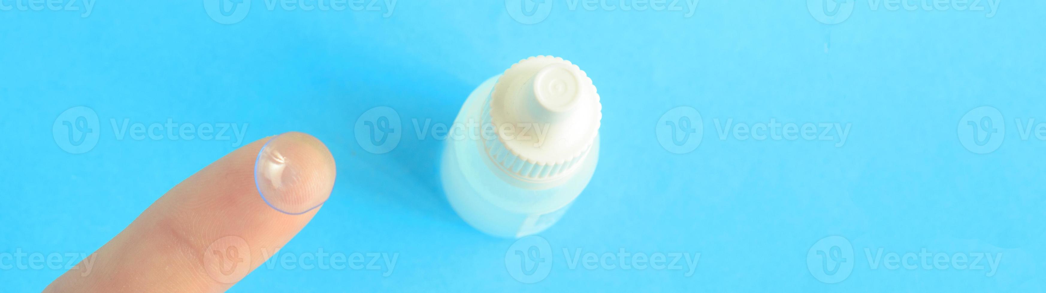 una lente de contacto en un dedo y una pequeña botella de gotas para los ojos en un fondo azul, espacio de copia foto