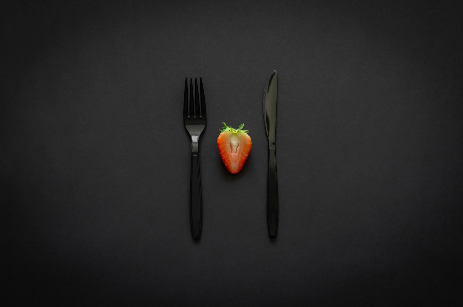 Cutting strawberry with fork and knife on dark background for minimalist flat lay black food concept. photo