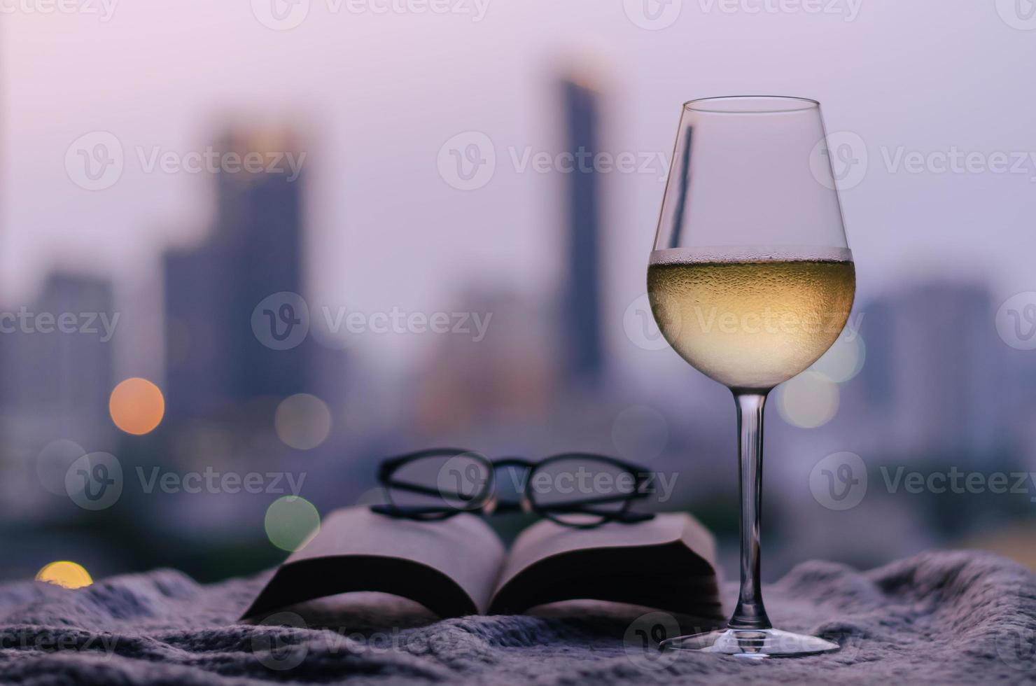 A glass of white wine with book and spectacles put on bed with colorful city bokeh lights background. Stay home concept. photo