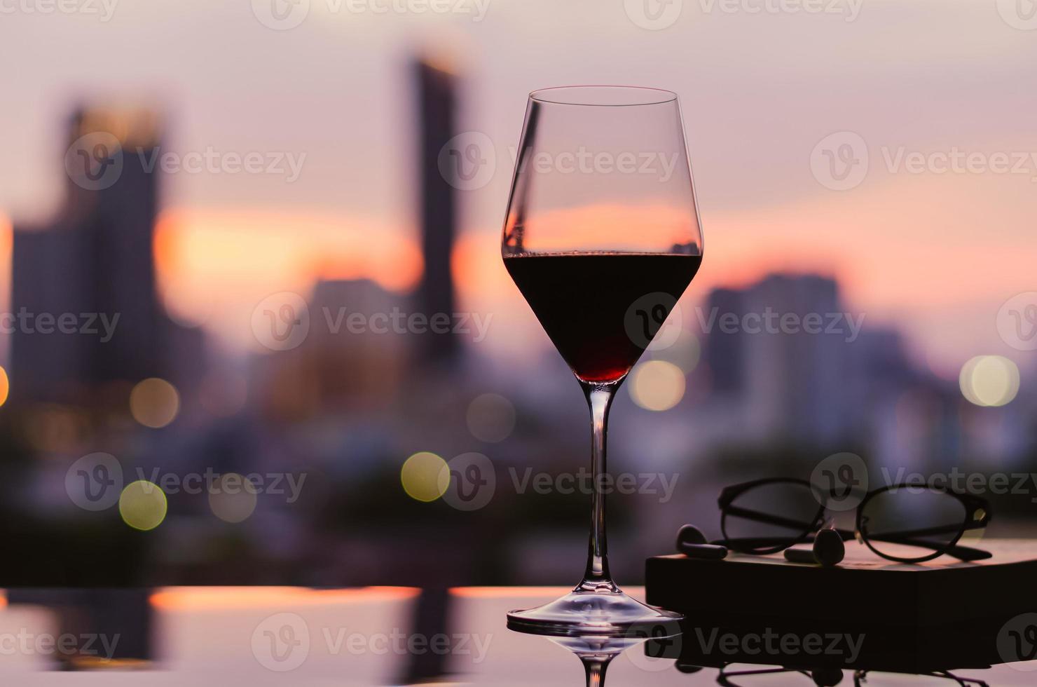 una copa de vino tinto con gafas y un auricular en un libro puesto en la mesa con el fondo de la ciudad. quedarse en casa concepto. foto